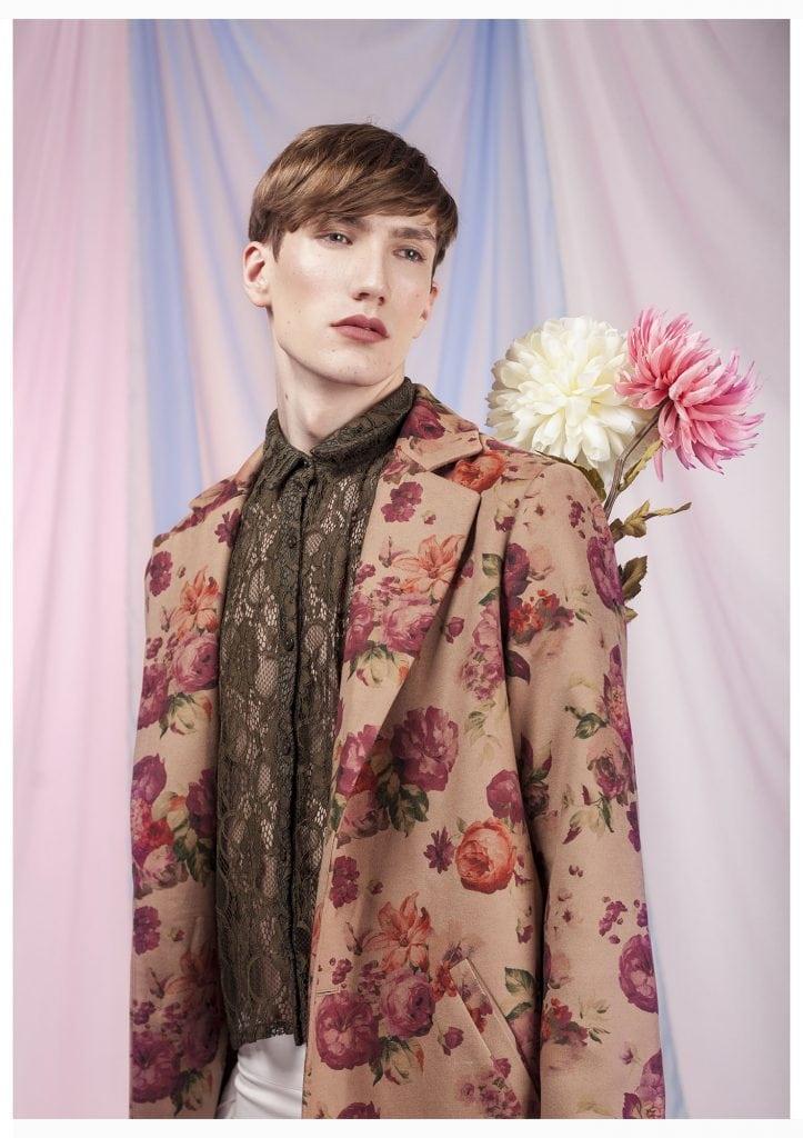 Image of a man in a floral suit jacket with a pair of flowers held behind his back, visible from above the left shoulder.