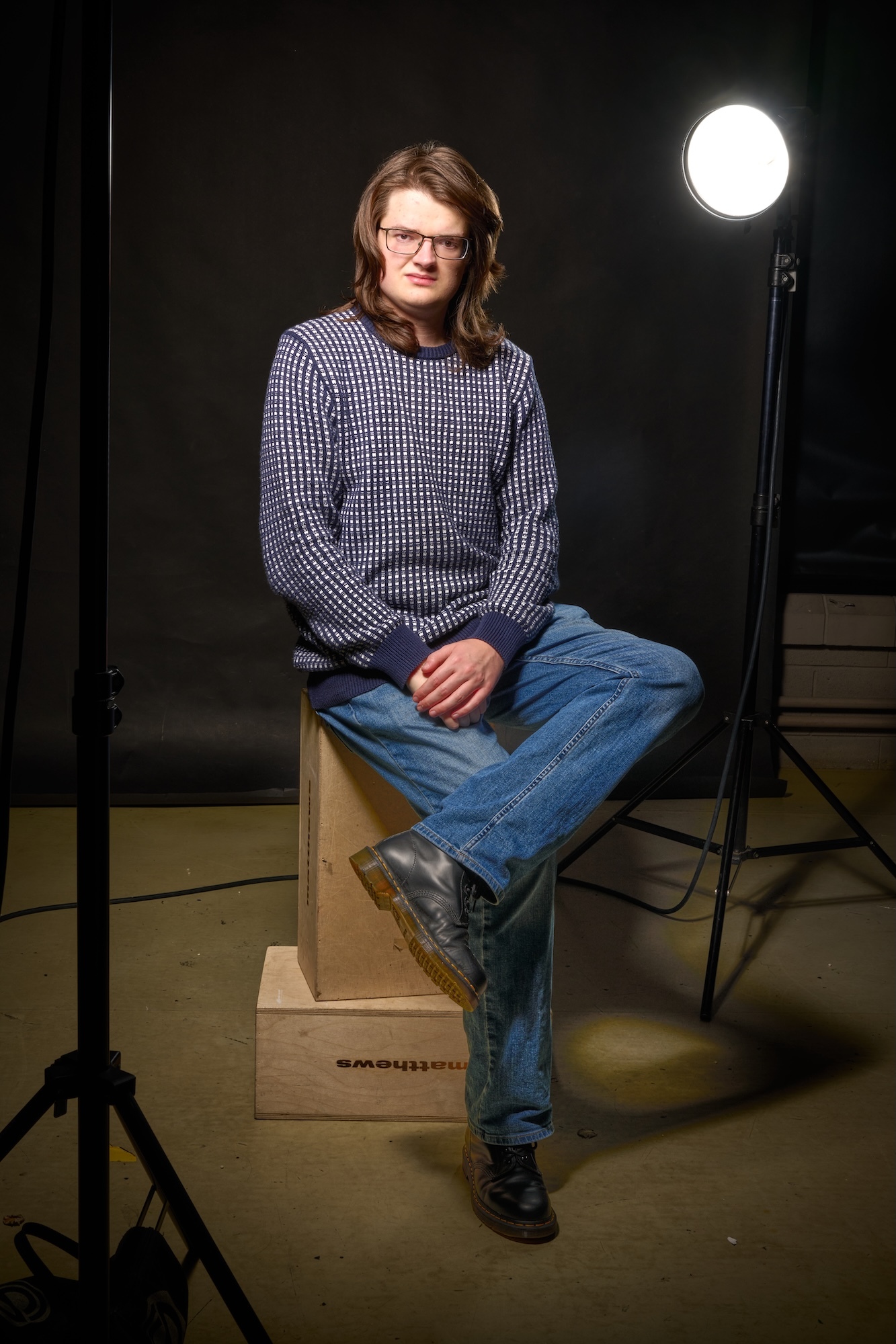 Portrait of BA (Hons) Photography graduate D Bowhay, sitting on wooden box in photography studio, wearing grey jumper and jeans with studio light visible.