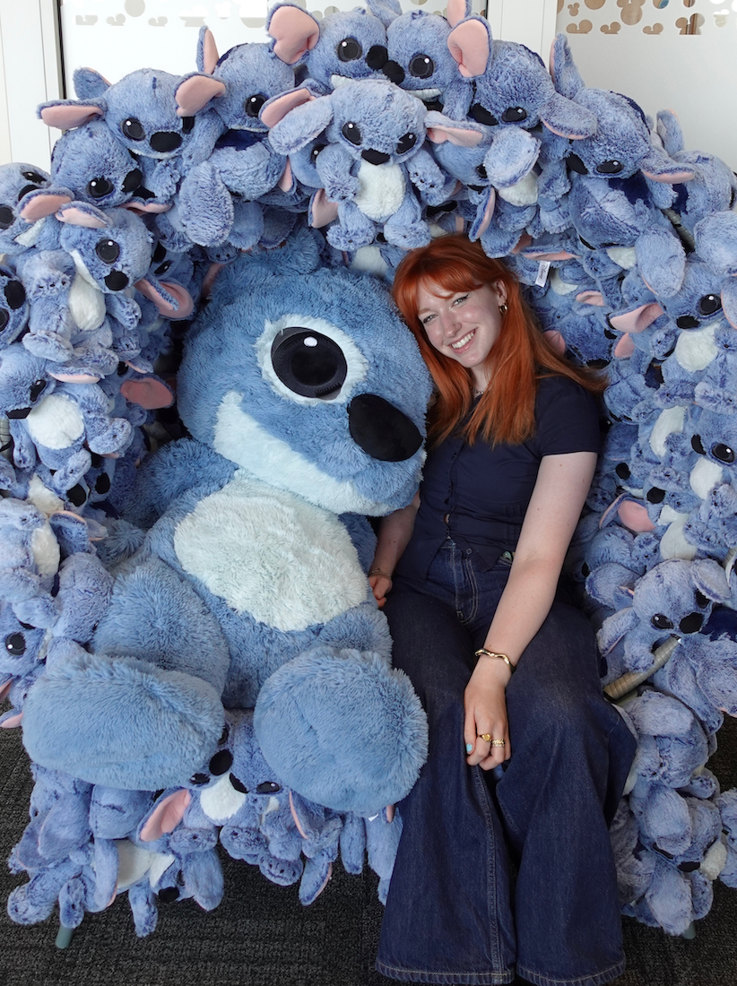 E Jarvis sits amongst dozens of plush Stitch toys covering wall behind her.