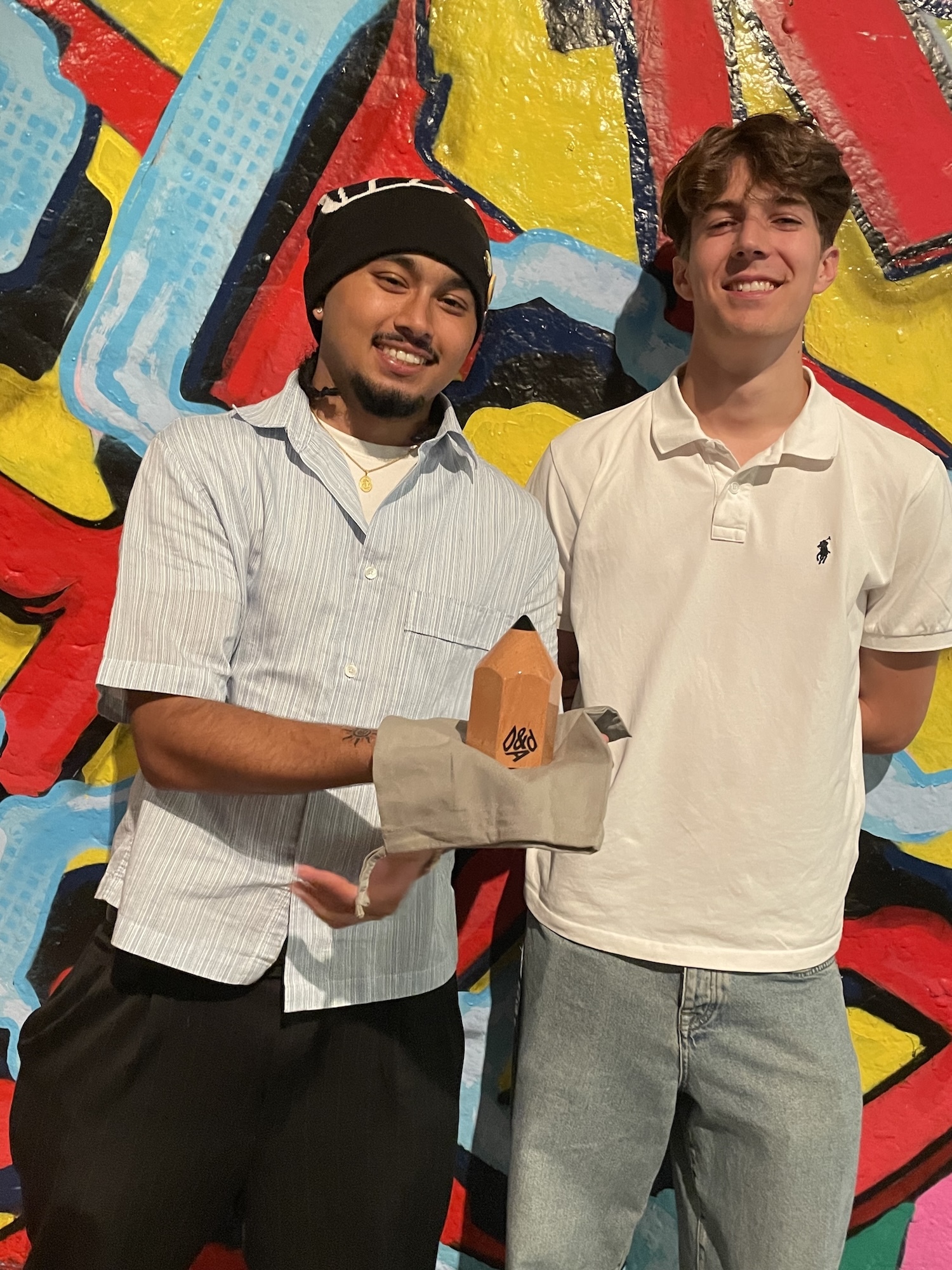 P Bhandari and B Courtenay standing together against vibrant graffiti wall with red, yellow, blue geometric shapes. One wears striped shirt and beanie, other in cream polo shirt and jeans.