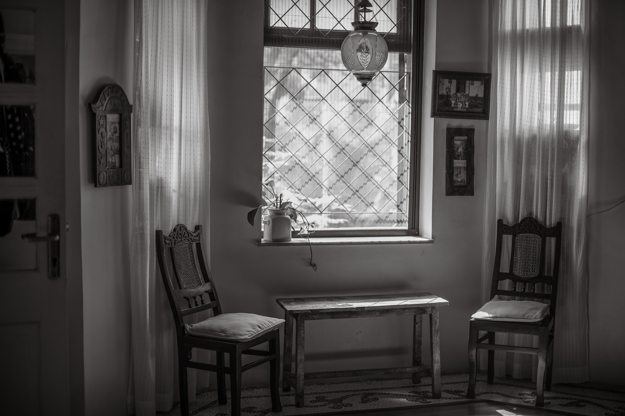 Black and white interior with two wooden chairs flanking small table beneath diamond-paned window with curtains and wall frames.