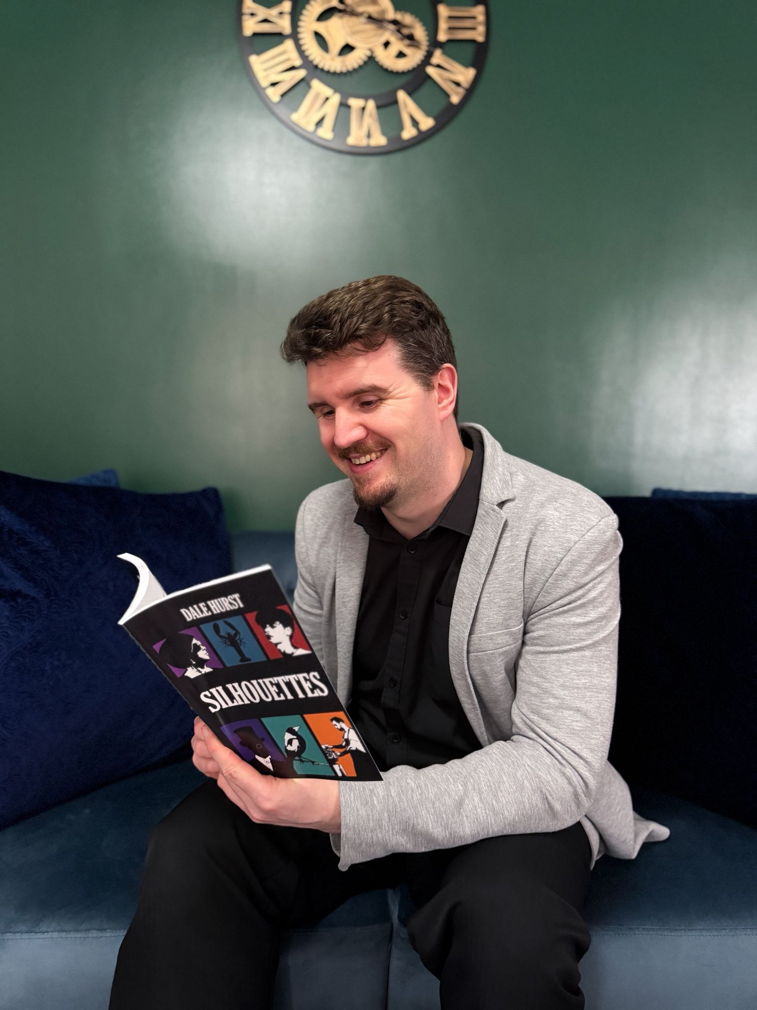 Man in grey blazer reading book titled "Silhouettes" on navy sofa. Large vintage clock on green wall behind.
