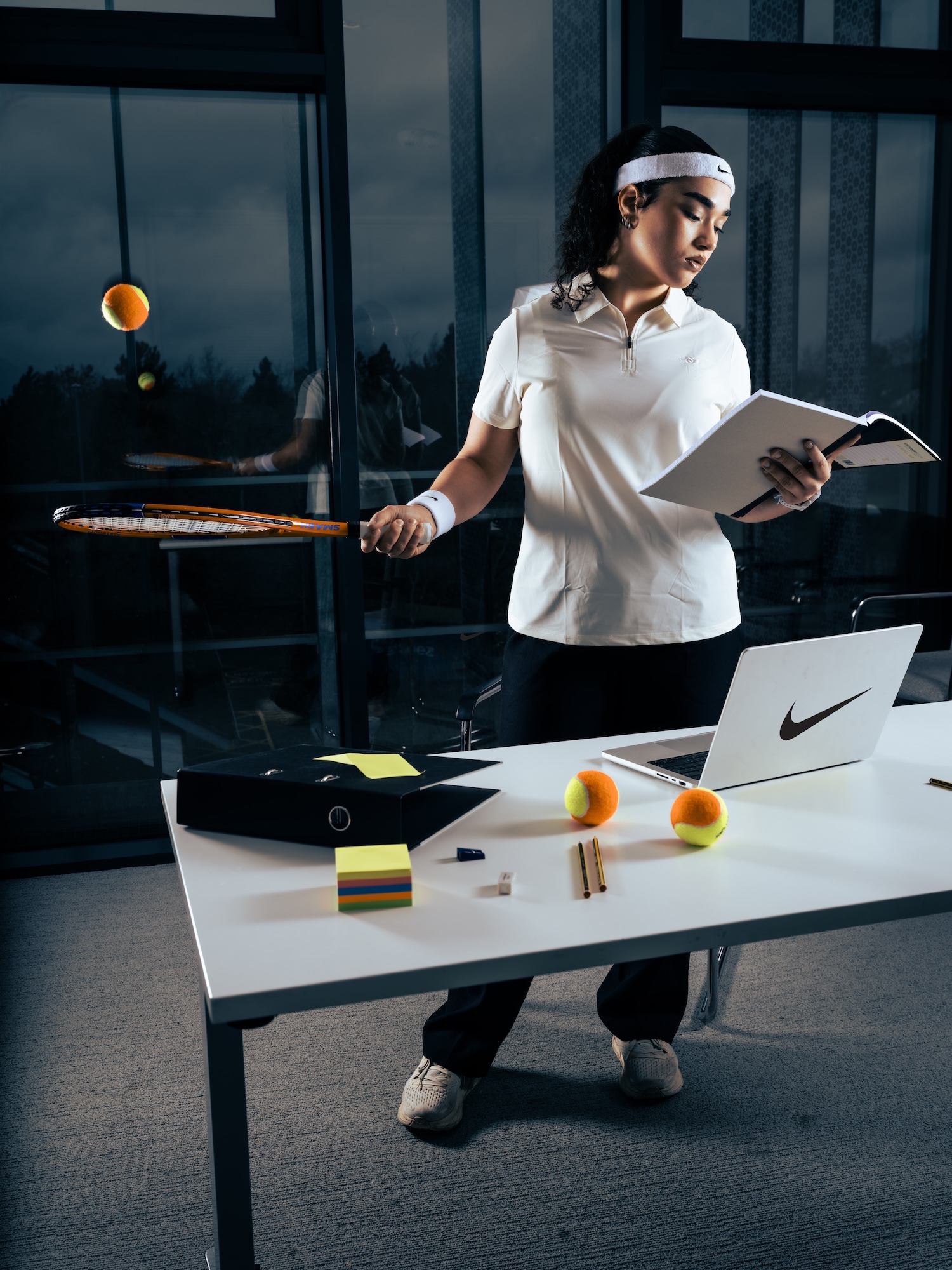 A young woman in a white polo shirt and headband stands at a table, holding a book in one hand and a tennis racket in the other. On the table, there is a laptop, a black box, sticky notes, and several tennis balls.