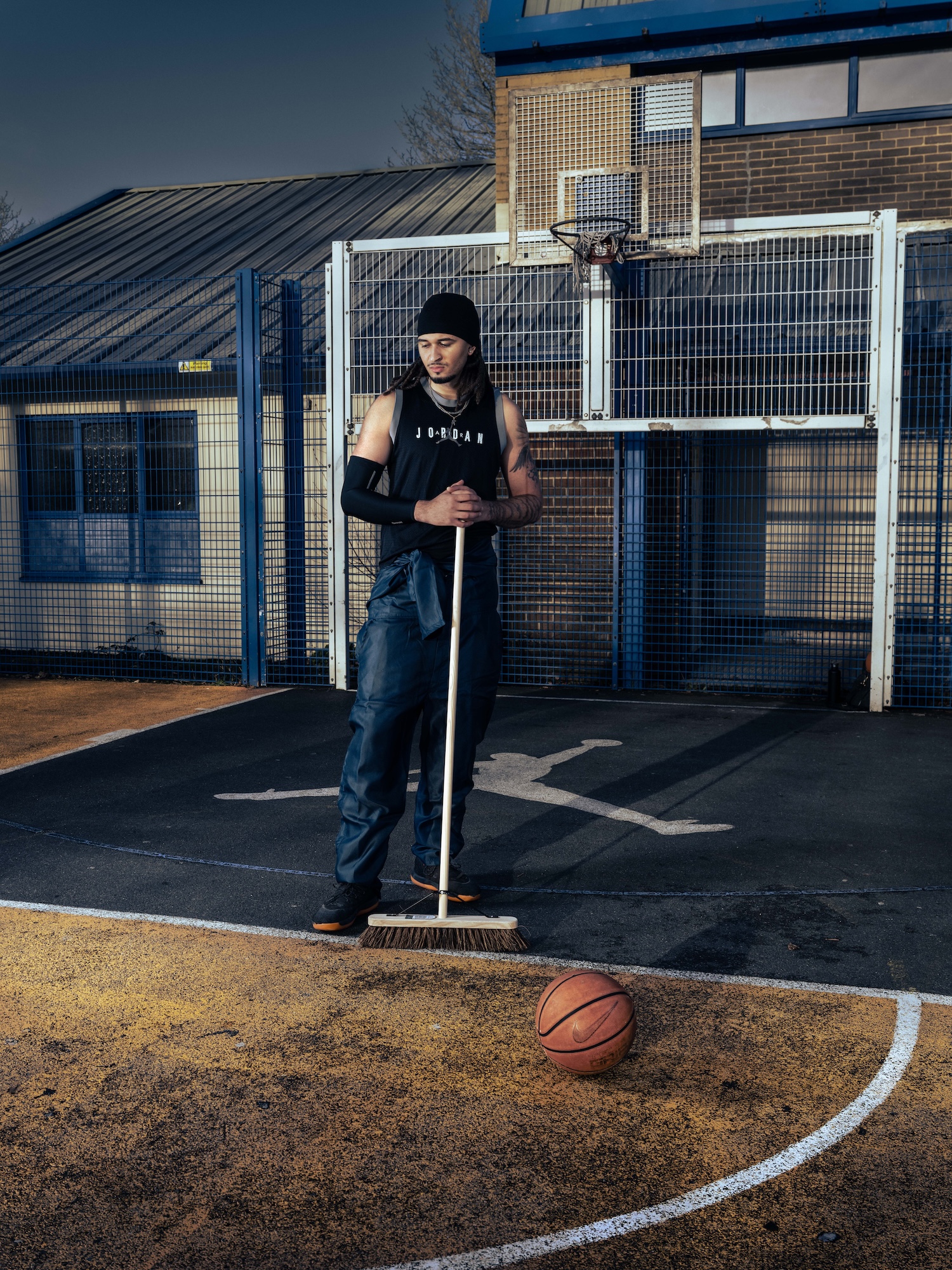 A man wearing a black sleeveless top and jeans stands on a basketball court, holding a broom. A basketball sits on the ground near his feet, and a building with large windows is visible in the background.