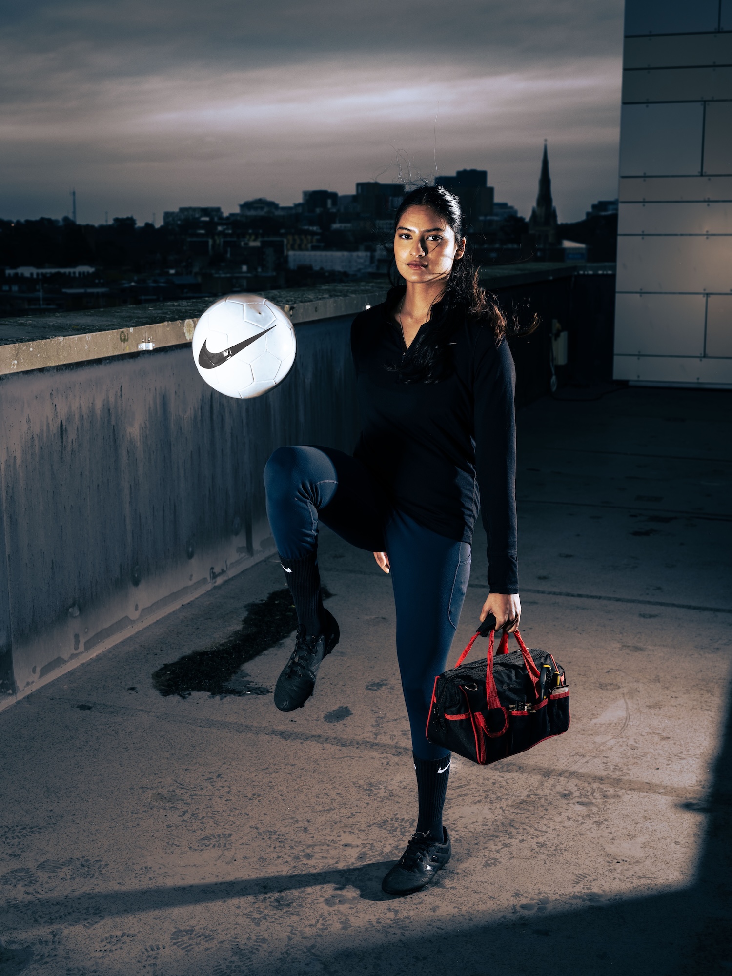 A young woman with long hair stands on a rooftop, balancing a Nike football on her knee. She wears a black long-sleeve top and navy leggings, holding a black and red sports bag in her right hand. The city skyline is visible in the background.