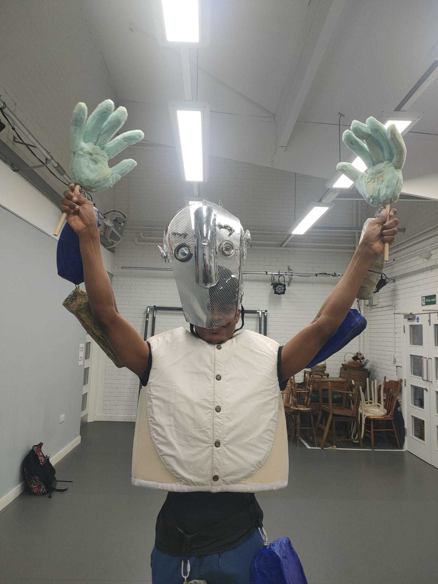 A BA (Hons) Dance student in partial costume wearing a mask/headdress. Their arms are raised and they are holding a large green hand in each hand.