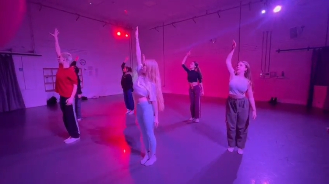 A small group of BA (Hons) Dance students rehearsing a routine in one of AUB's dance studios. They each have one arm pointing upwards, standing under red and purple lighting.