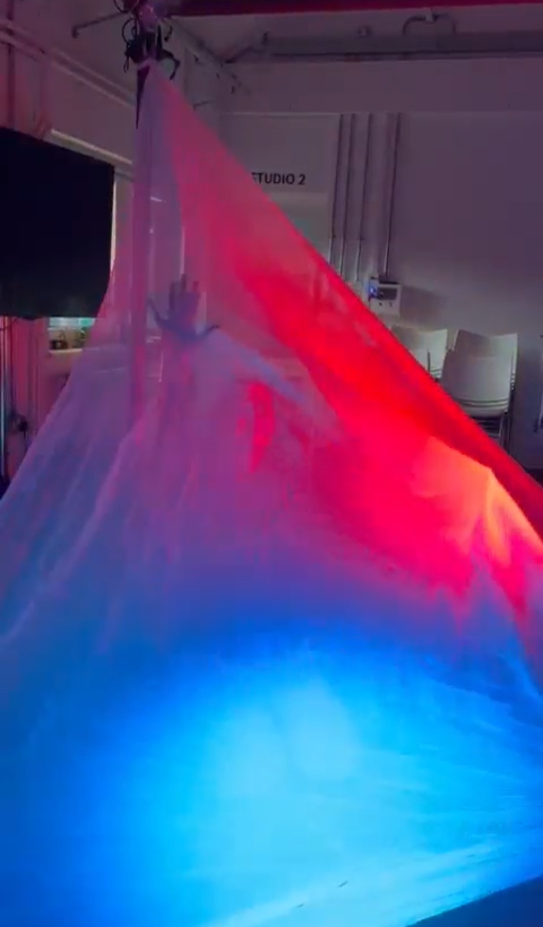 A BA (Hons) Dance student rehearsing. They are visible underneath a sheet covering them. Red and blue lights reflect off the sheet.