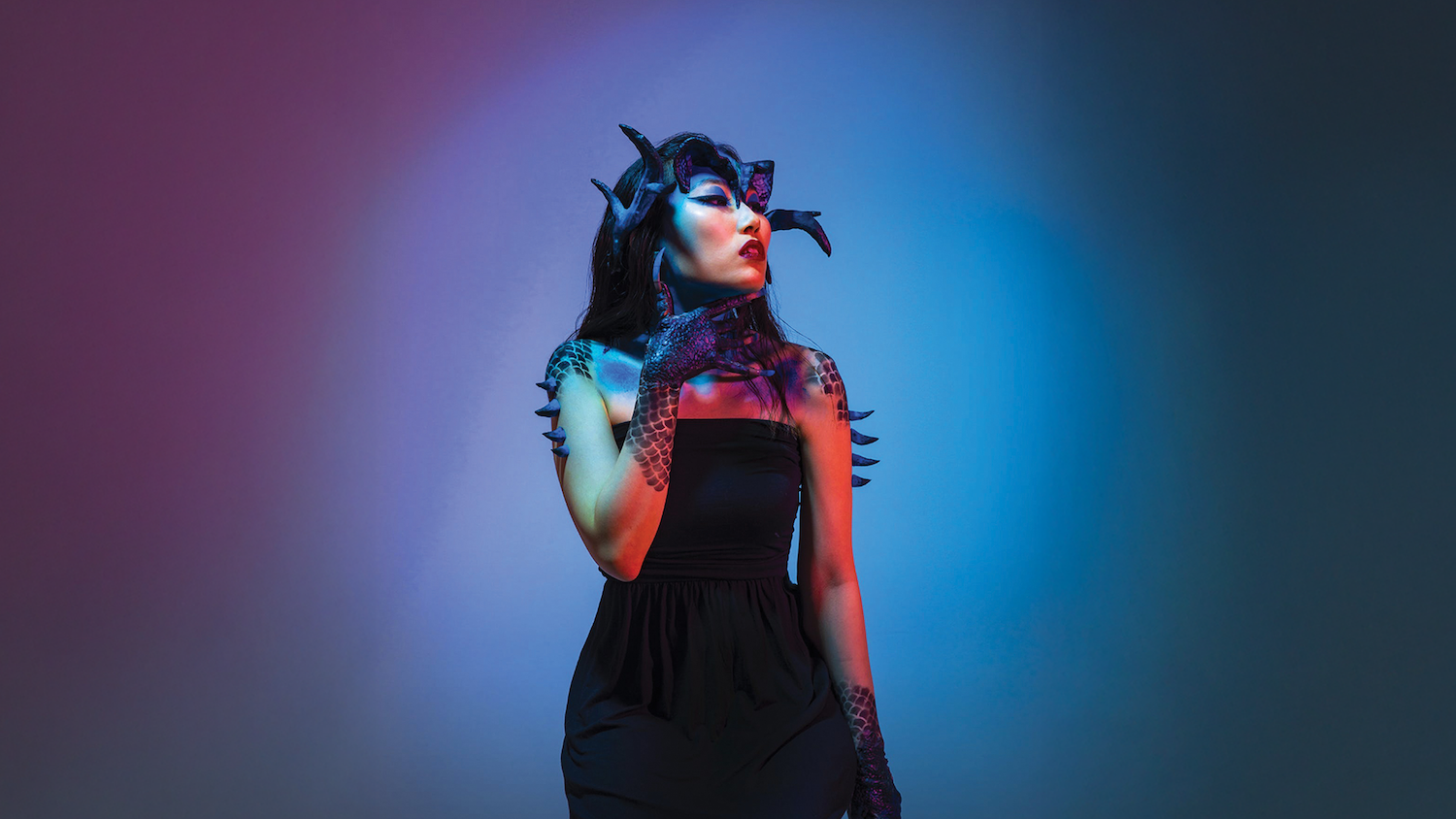 Woman in black dress with horned headpiece against purple and blue gradient background lighting.