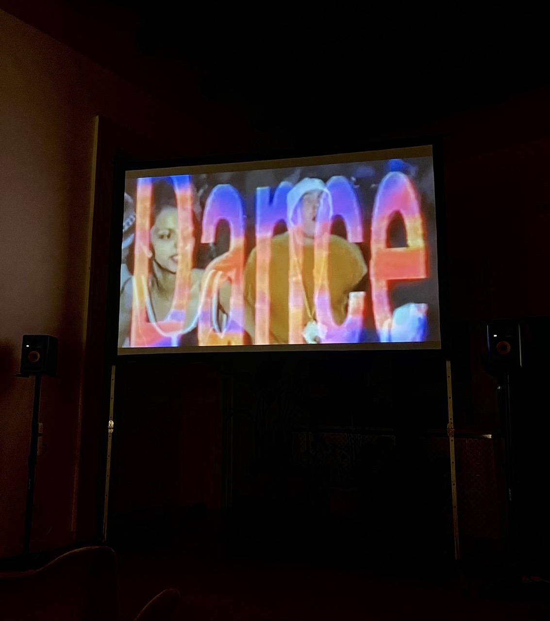 Still shot from RECOMMENDED DOSAGE in a dark room, screen shows VHS footage with 'DANCE' in blue, pink and orange text with people raving behind.