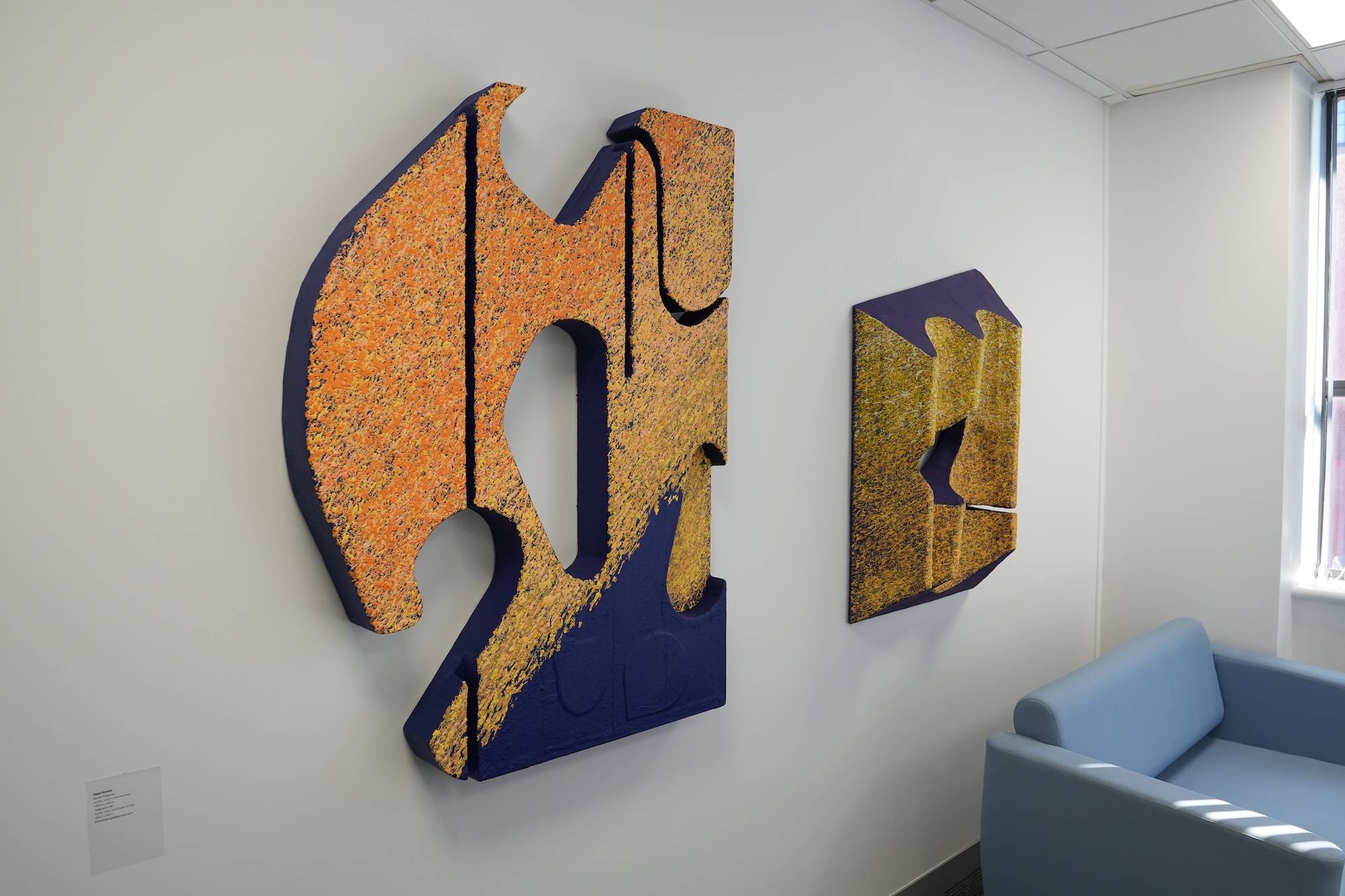 Two abstract wall sculptures in orange, gold and dark blue hang on a white office wall beside a light blue sofa and a window.