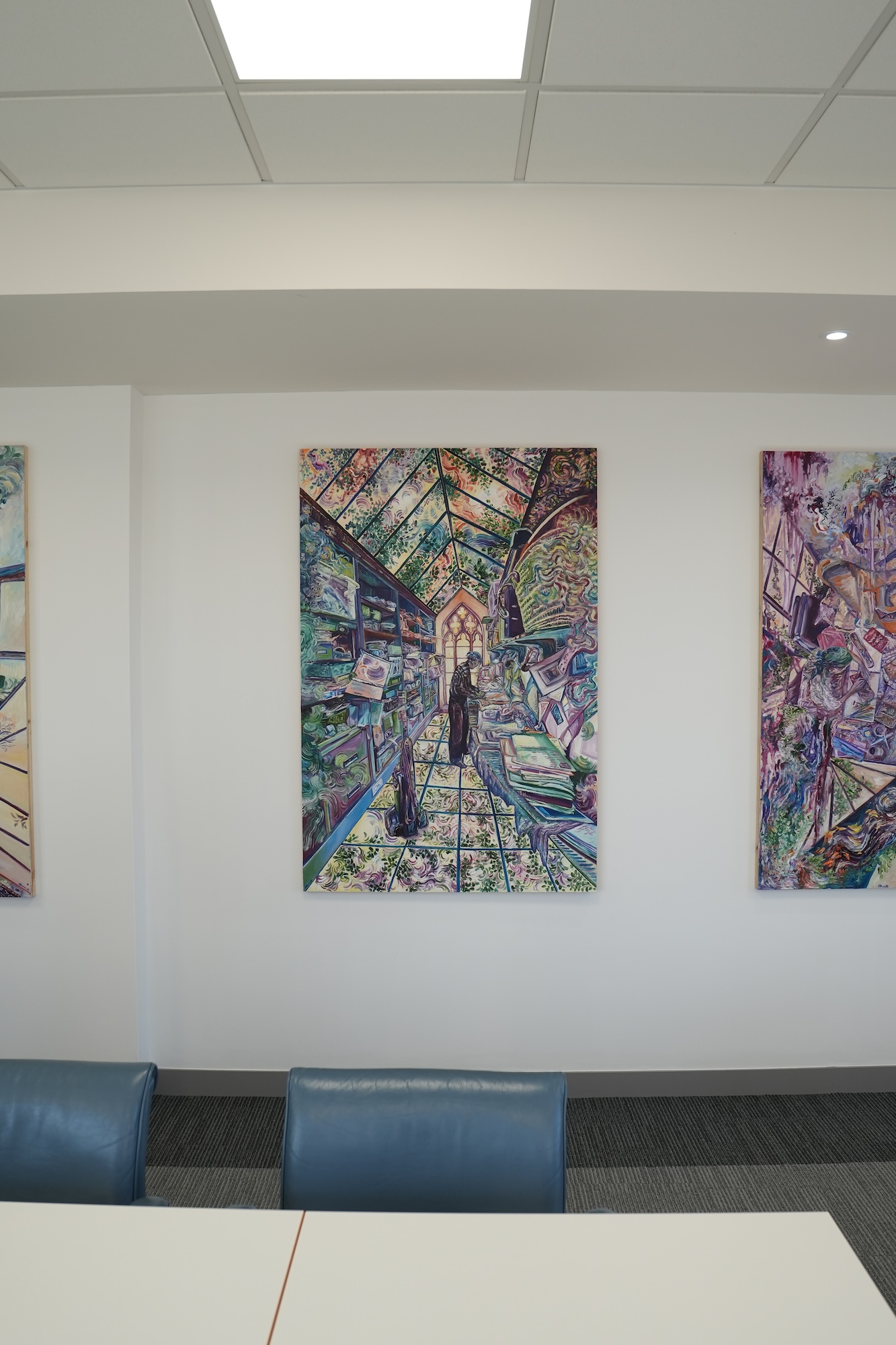Three colourful abstract paintings hang on a white wall above blue chairs and a white table in a modern interior.