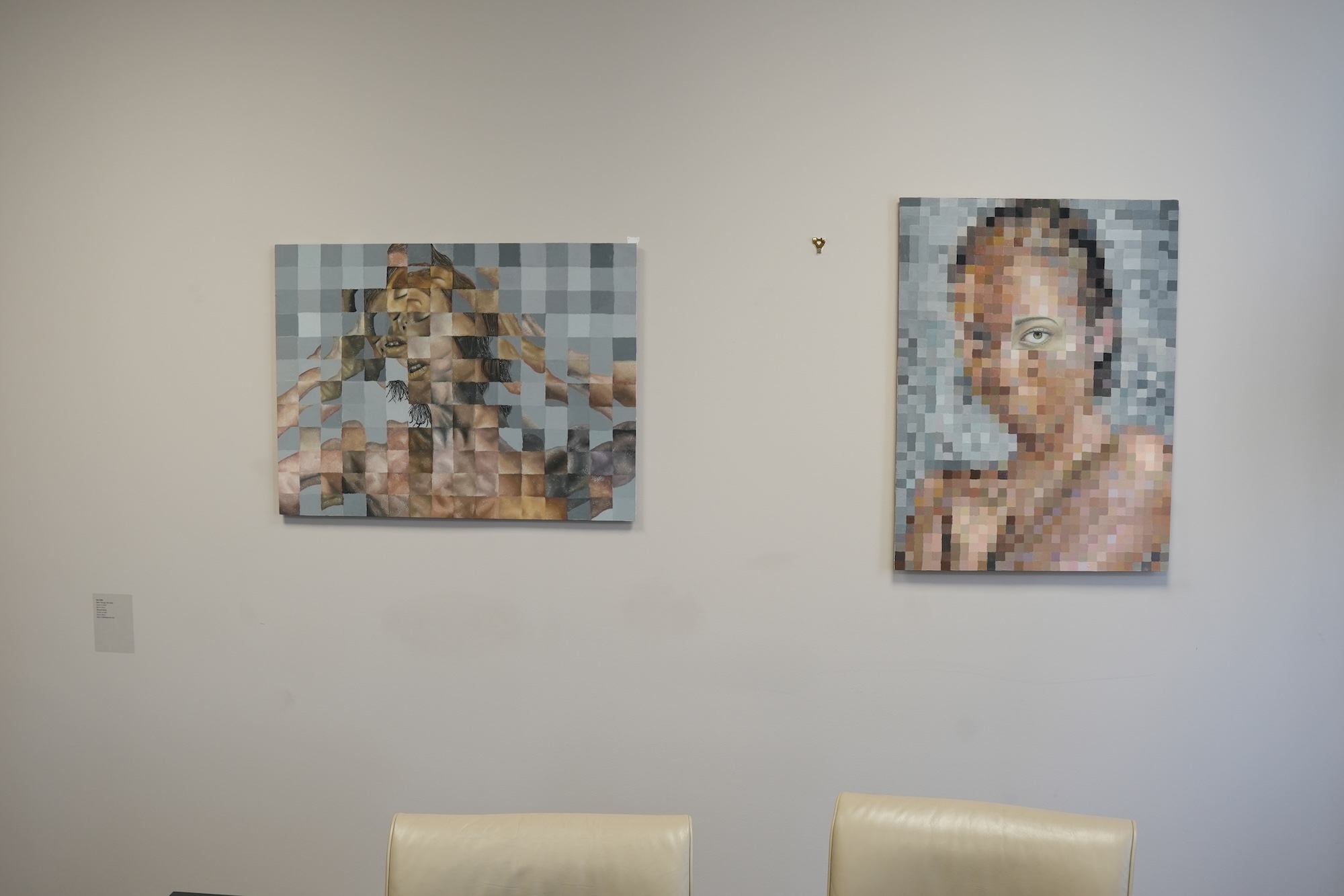 Two pixelated portrait artworks hang on a plain white wall above cream chairs.