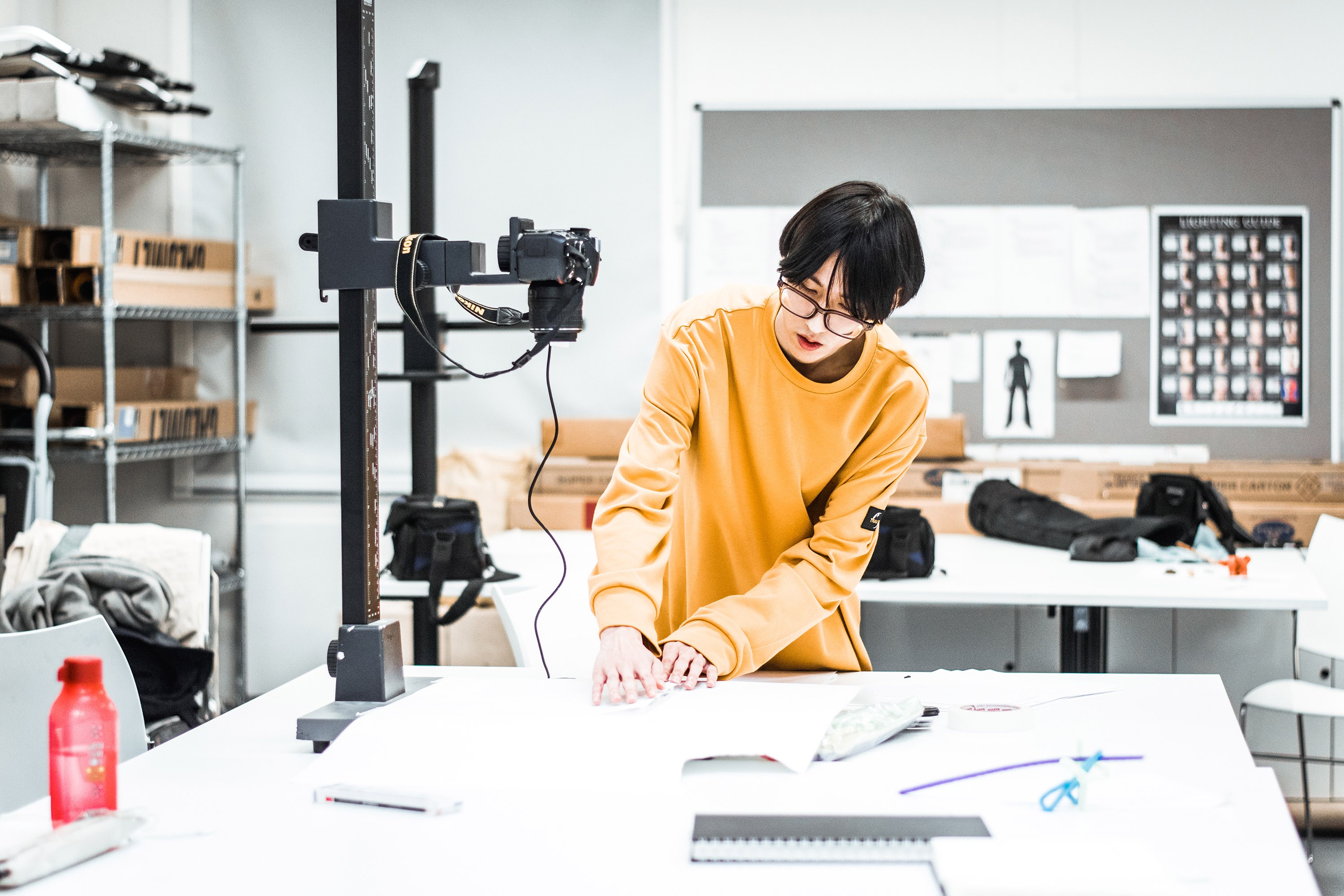A student working at a table in a studio, lining something up to prepare to take an overhead photo. A camera is fixed to a rig on the left-hand side pointing down towards the table.