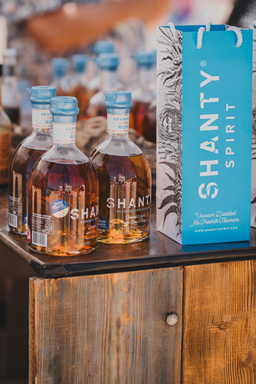 Bottles with orange rum and light blue cap and bag next to them reading 'Shanty Spirit', on a wooden stand