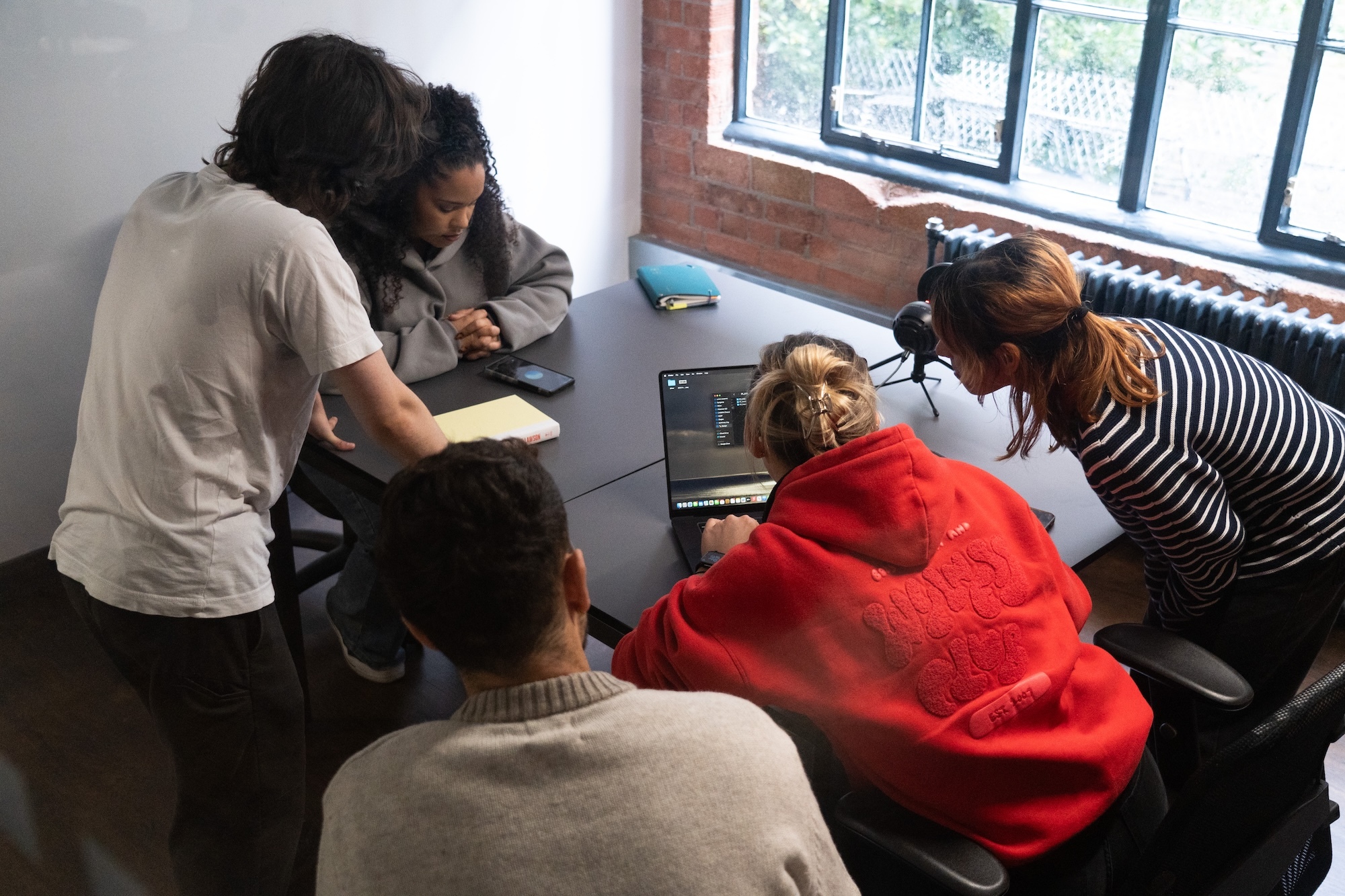 A team of five students gathering around a single laptop in a well-lit classroom space.