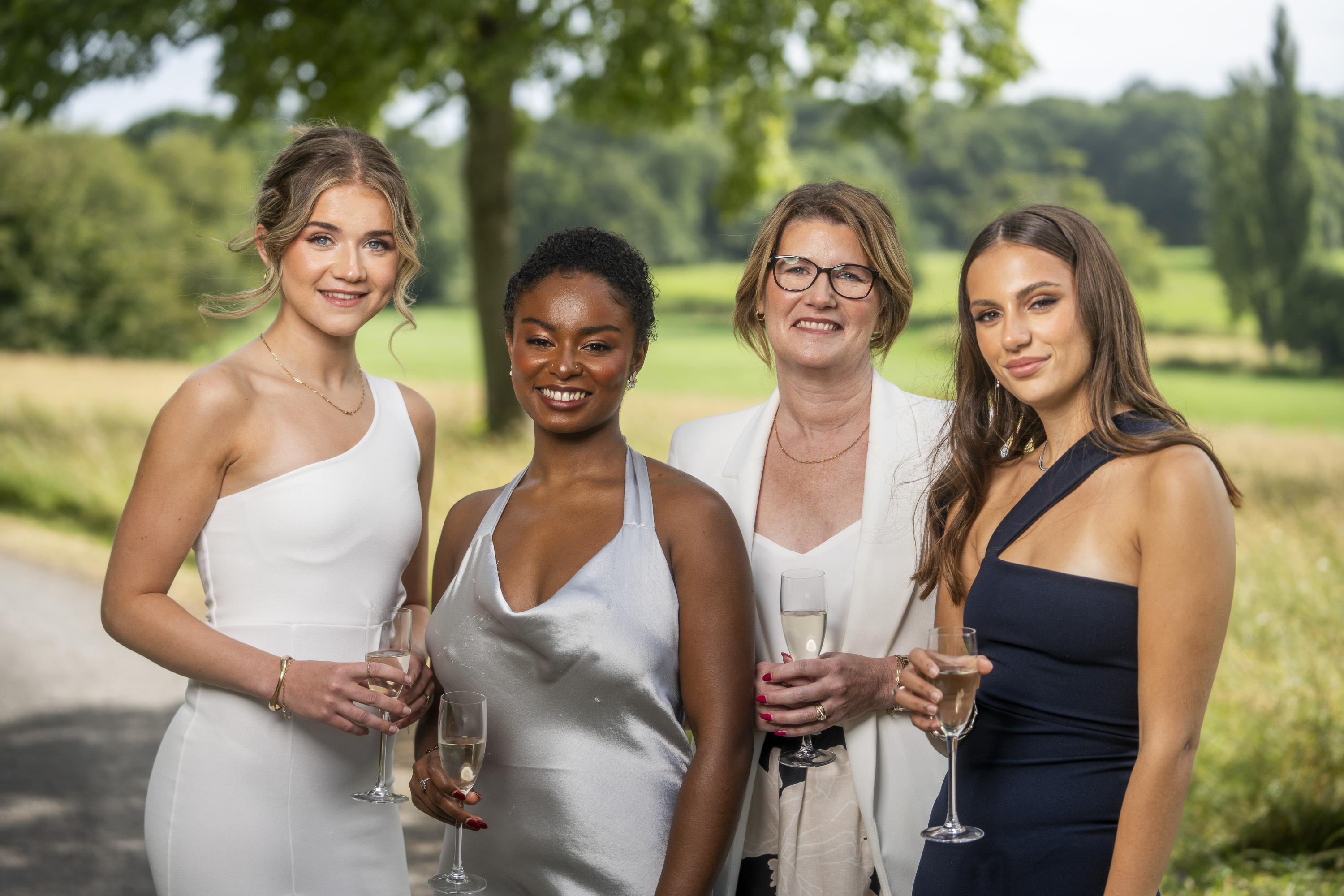Four women in formal dresses holding champagne glasses, standing outdoors with green countryside and trees in background.