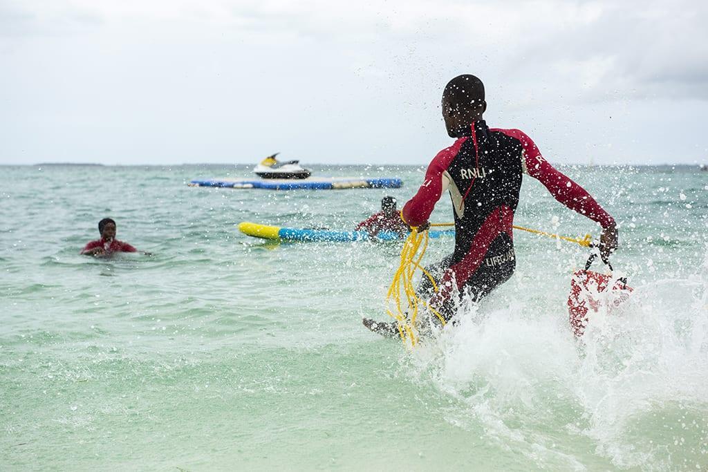 Person wearing an RNLI wetsuit runs into the sea towards two other people.