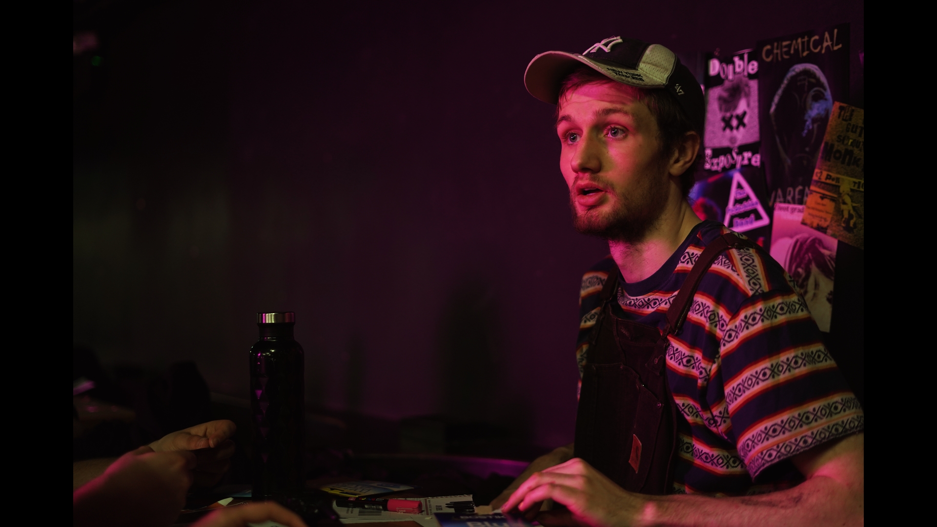 A young man with a beard and a cap stands behind a counter, wearing a striped shirt with a black overall. He is engaged in conversation, with various posters and a water bottle visible on the surface in front of him. The setting is dimly lit with purple and pink hues.