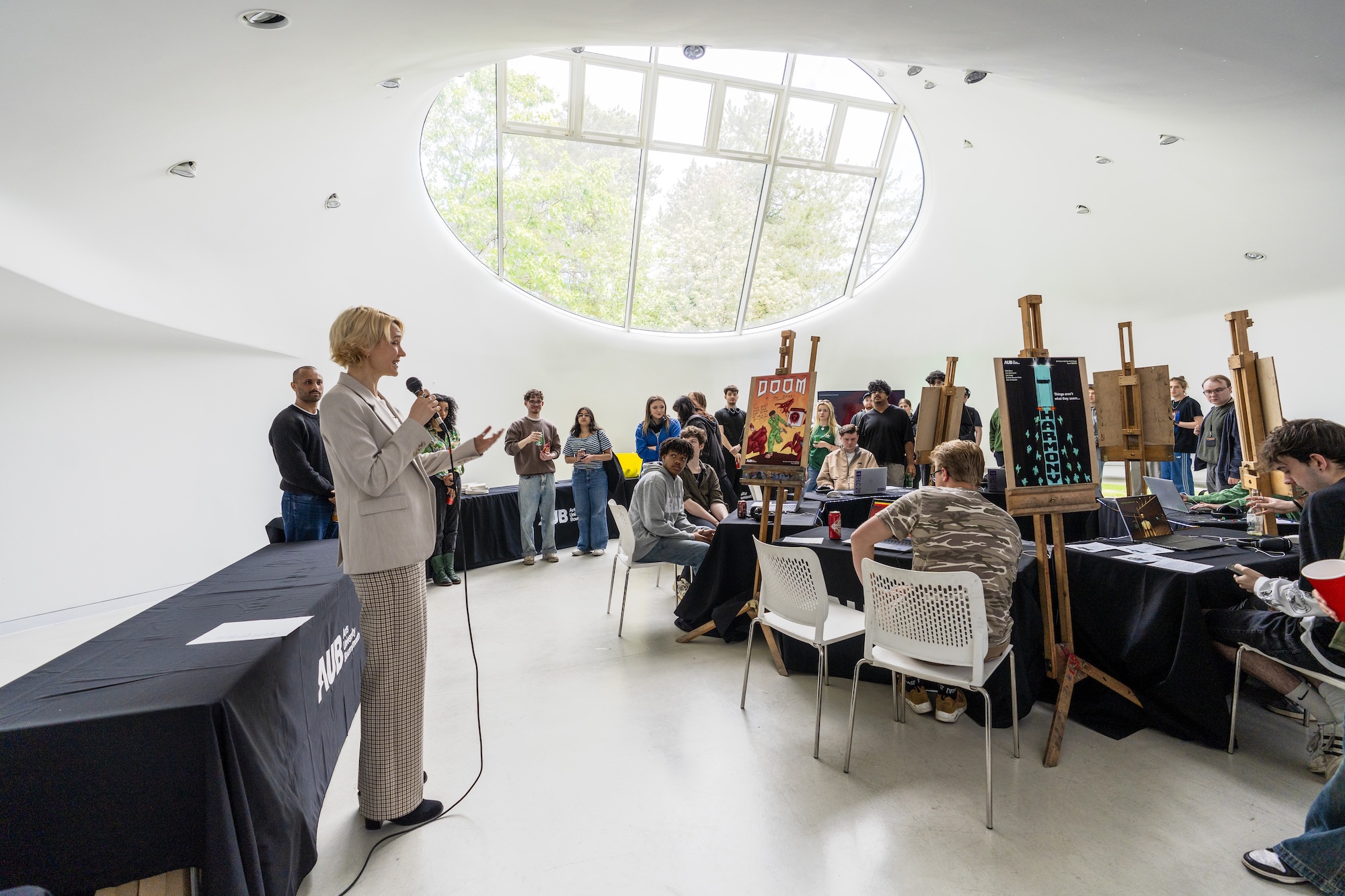 Dr A. Potemska stands with a microphone addressing a group of Arts University Bournemouth students and staff seated at tables at a games art and design exhibition in The Drawing Studio, AUB Campus.