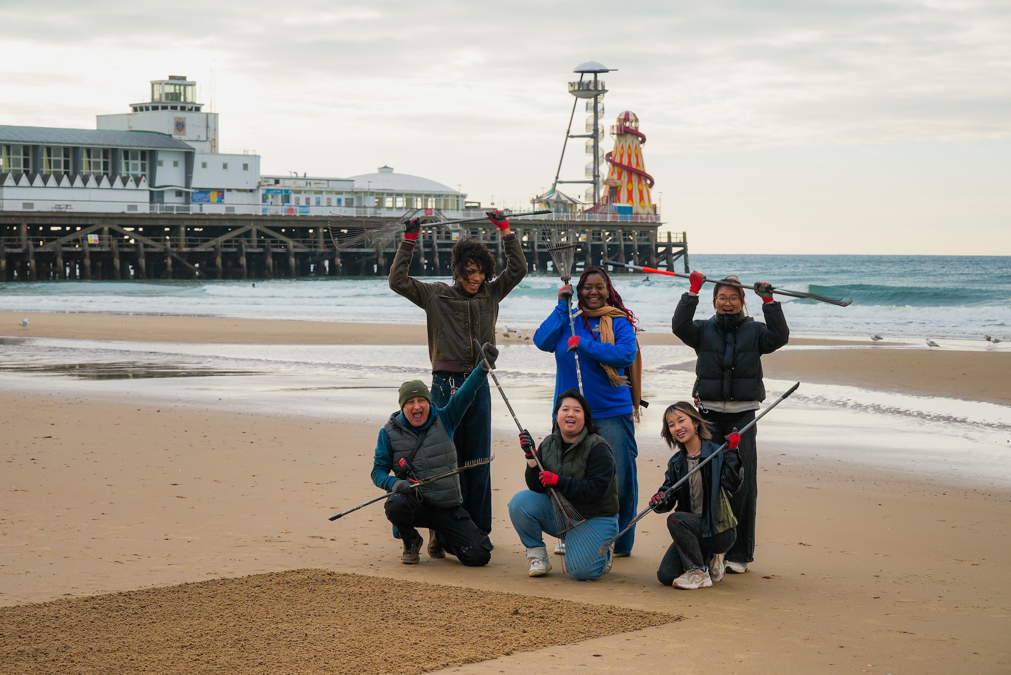 Six people posing (three standing, three kneeling) on Bournemouth Beach holding rakes. Bournemouth Pier is visible in the background.