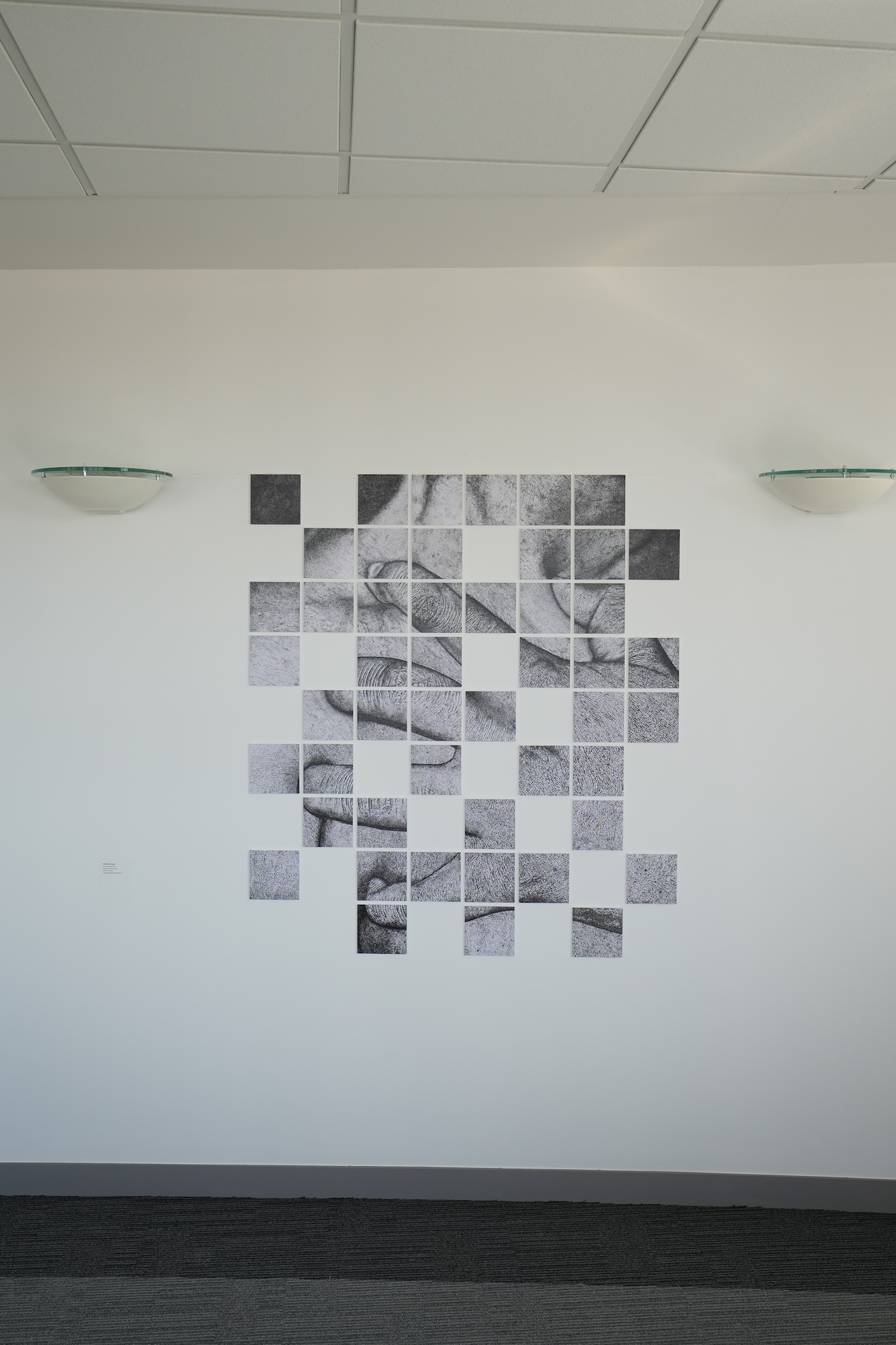 A monochrome square-tile wall artwork arranged in a grid on a white interior wall, with two glass light fittings mounted on either side and a tiled ceiling above.