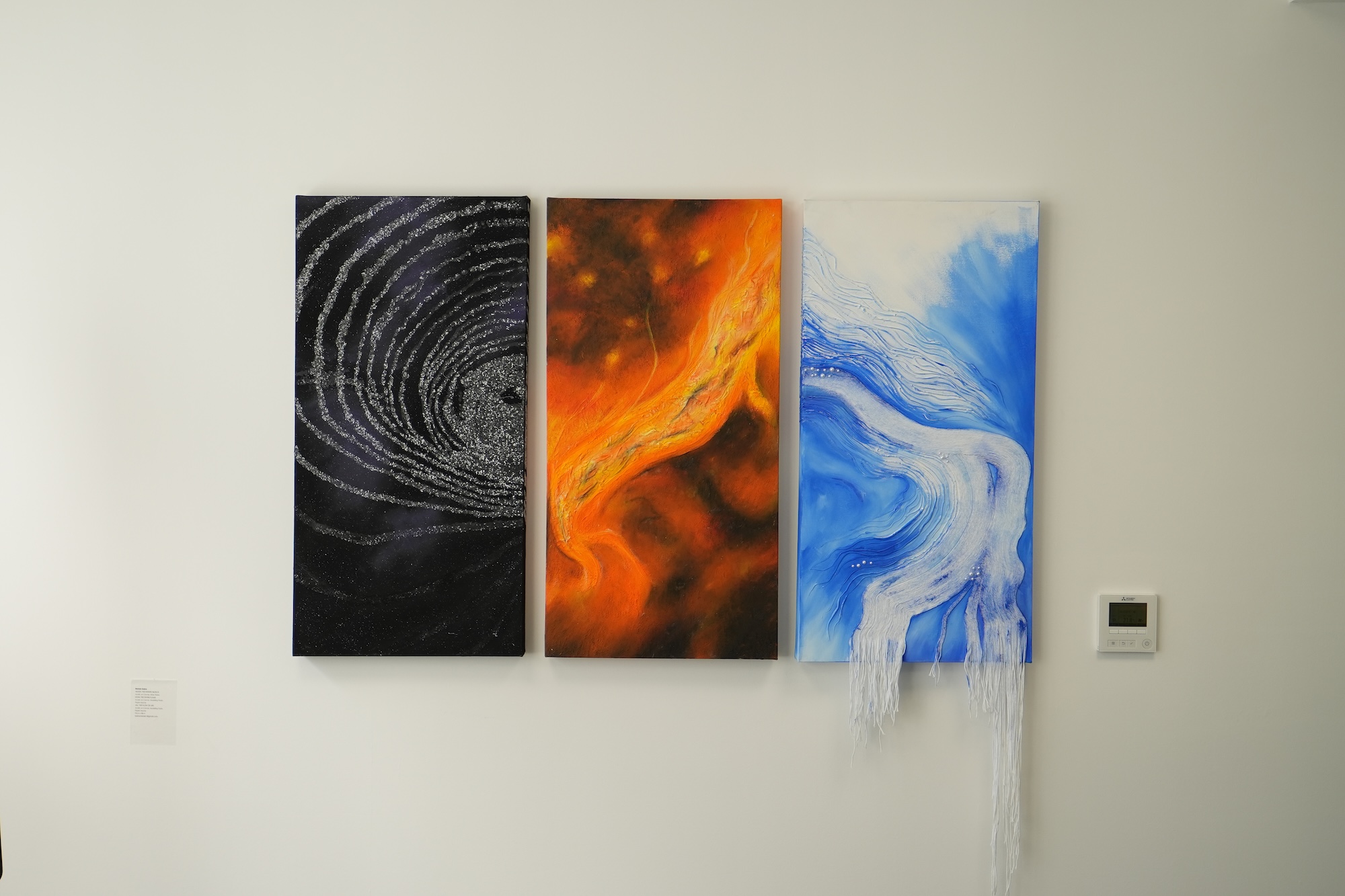 Three vertical abstract paintings hang on a white wall, showing black-and-grey swirls, orange flames and blue-white waves, with a small control panel to the right.