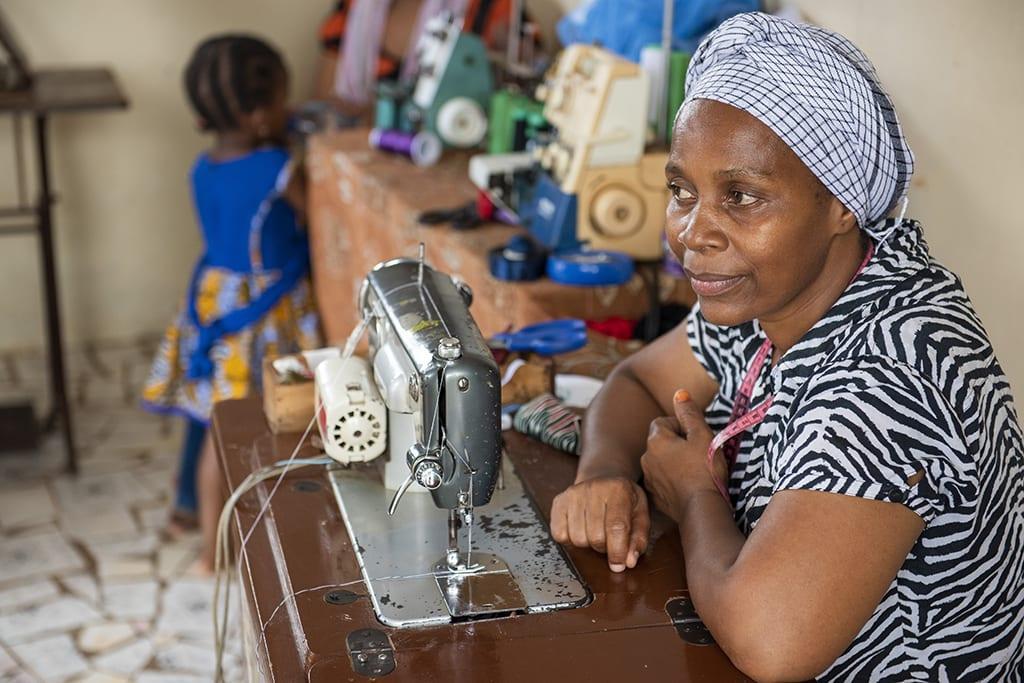 Woman sits by a sewing machine, looking away from the camera, in a living room. A child with their back to the camera stands in the corner.