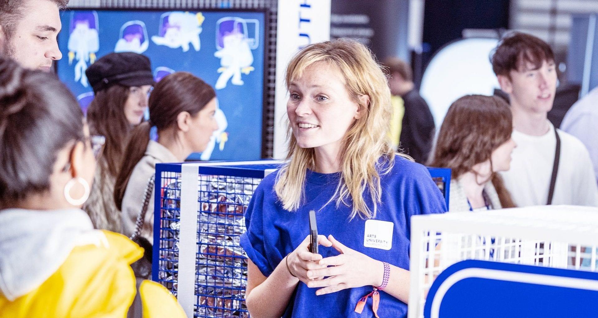 A busy image focussing on a female student ambassador in a blue, AUB branded t-shirt talking to a group of potential students at a careers fair