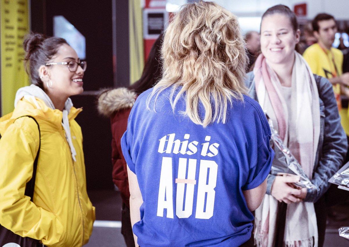 Student ambassador speaking with three other people. Their back is to the audience, they are wearing a T-shirt with 'This is AUB' printed on the back.
