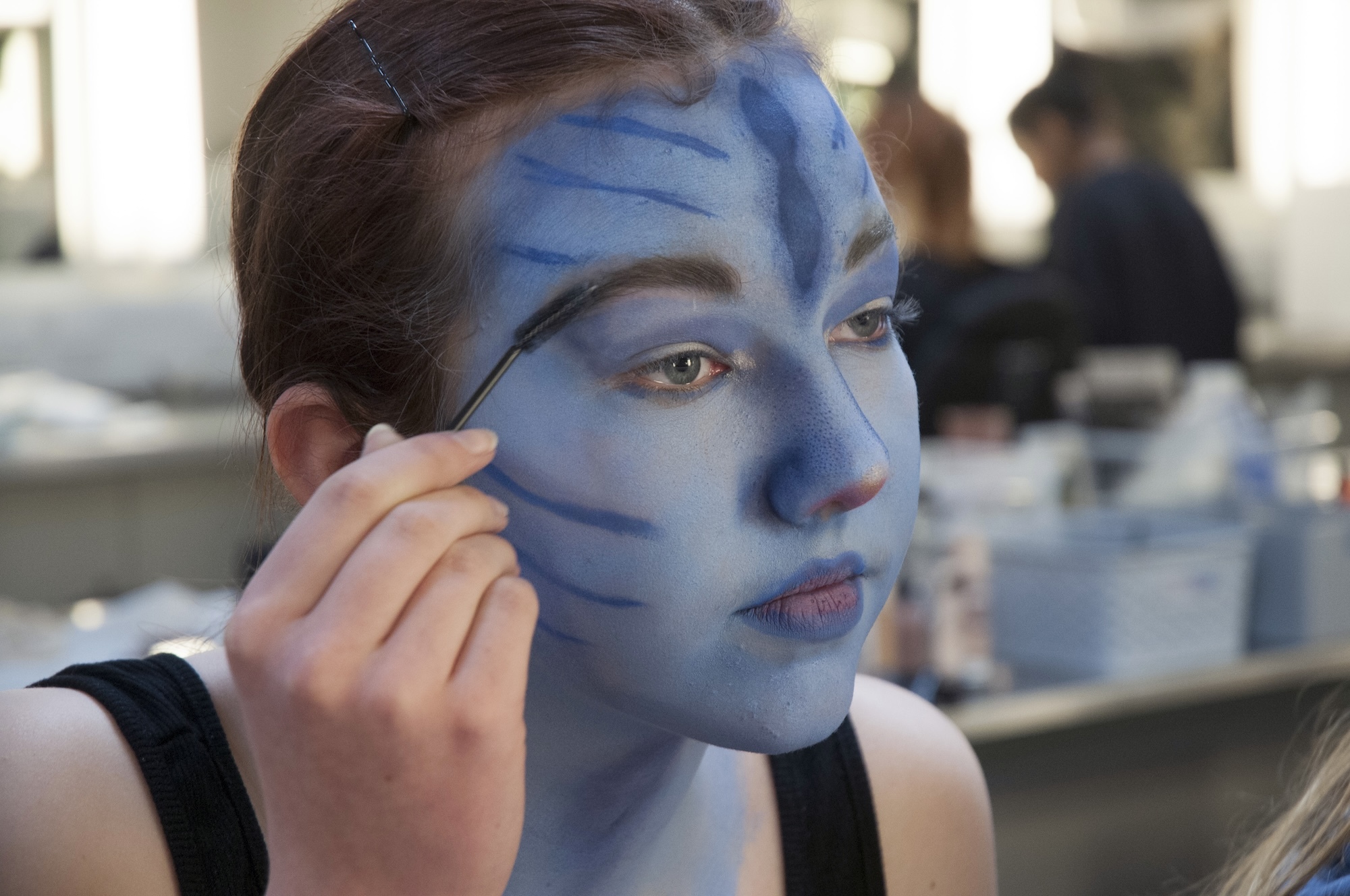Person applying blue face paint with black geometric lines using a brush, looking in mirror