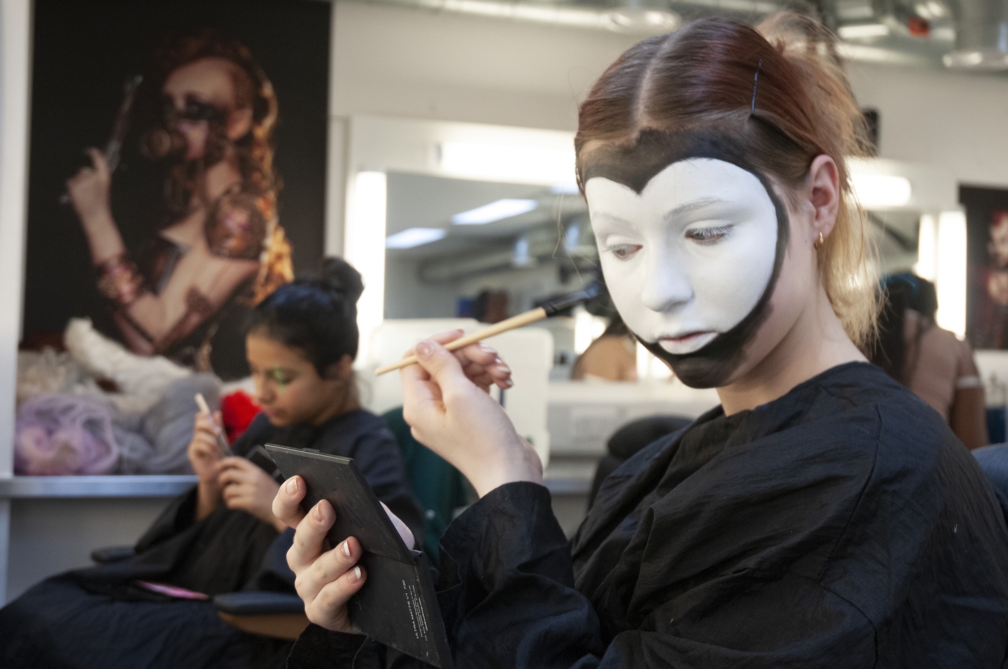 Person in black clothing wearing white theatrical mask applies makeup with brush whilst looking in mirror in busy salon