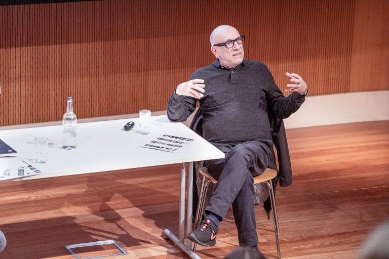 B Clarke sitting at one end of a table and speaking at an event.
