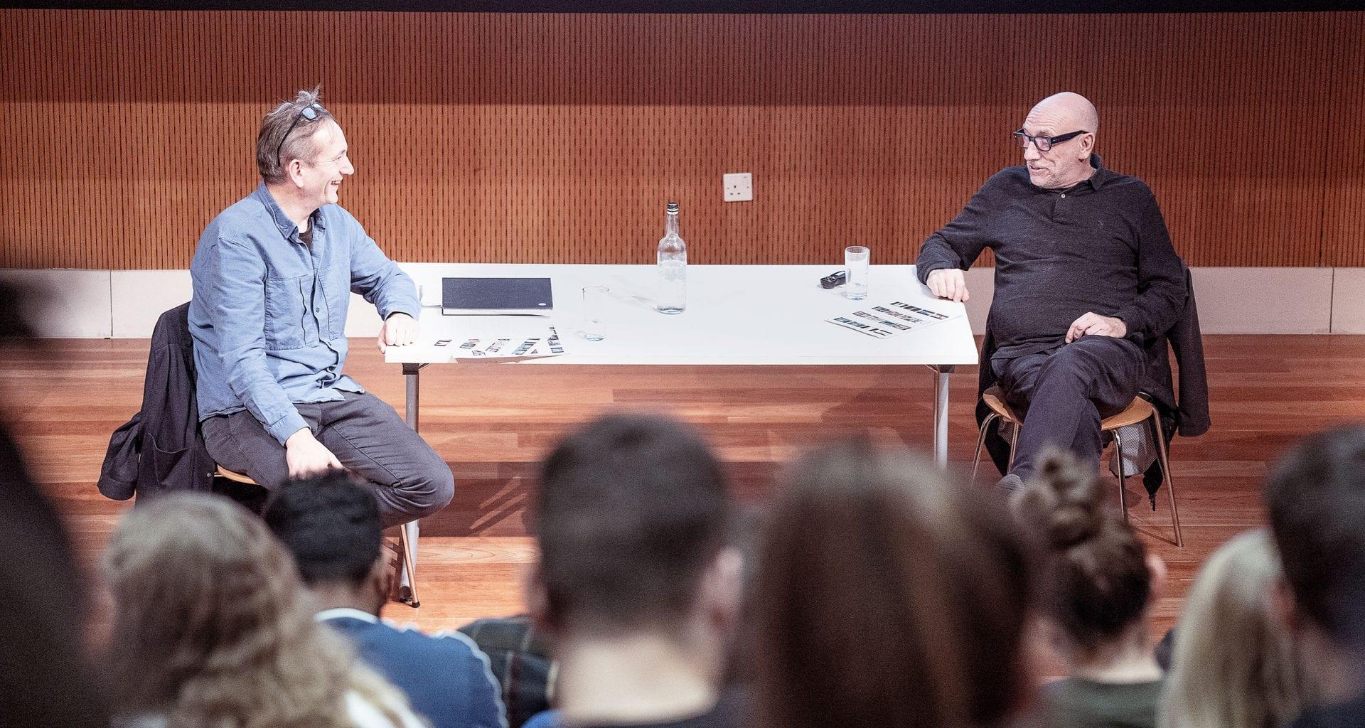 Two people sit at either end of a long table on stage and talk in front of an audience.