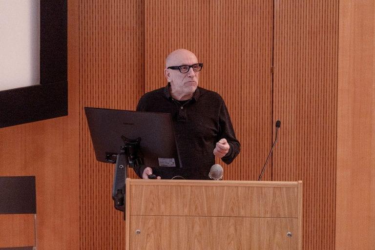 B Clarke standing behind a lectern and monitor delivering a lecture.