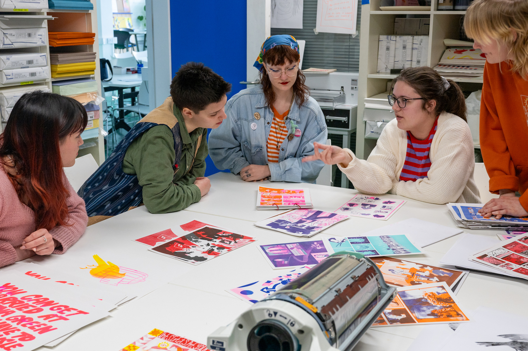 A group of students in colourful clothing looking at colourful illustrations on the table in a studio.