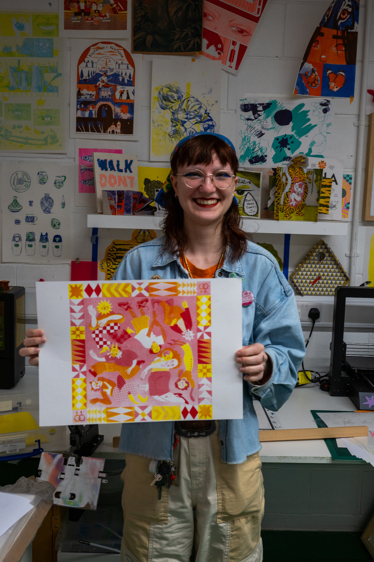 Artist holding up a colourful illustration, red, orange, yellow and pink, in a studio with artwork on the walls.