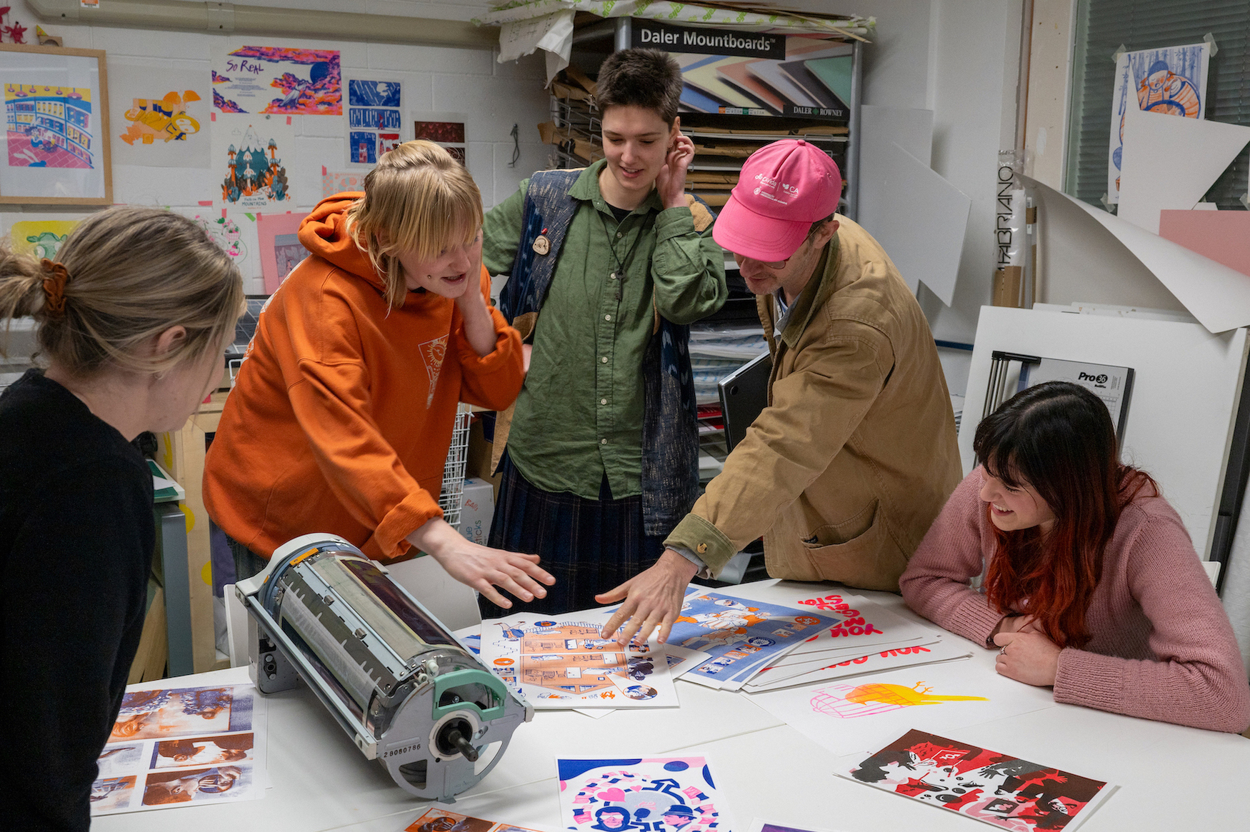 A group of students in colourful clothing looking at an illustration on the table in a studio, surrounded by other colourful artworks.