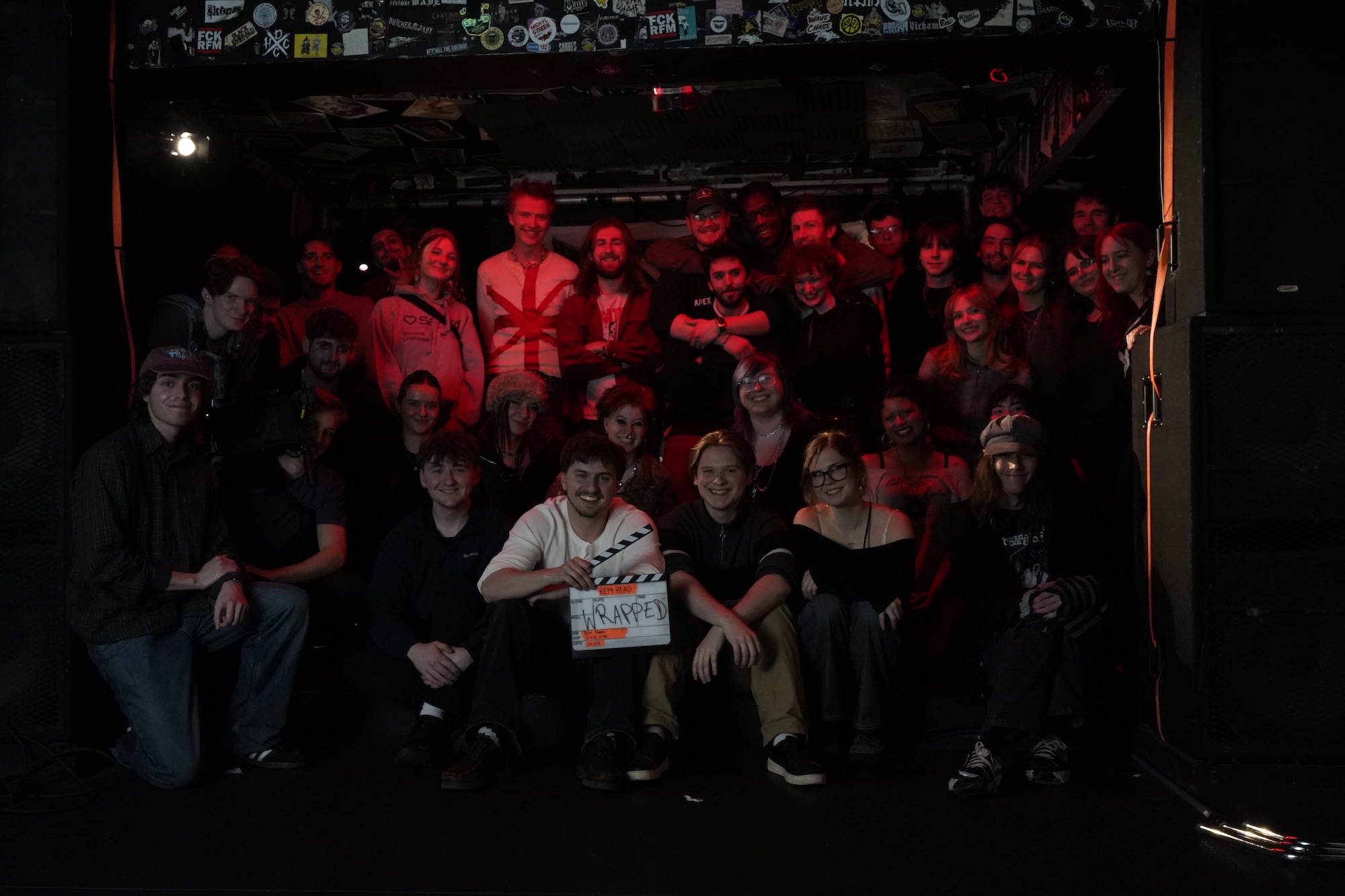 A large group of approximately 40 people gathered in a dimly lit venue. The front row features individuals holding a clapboard, while others stand and sit behind, with stickers visible on the walls in the background.