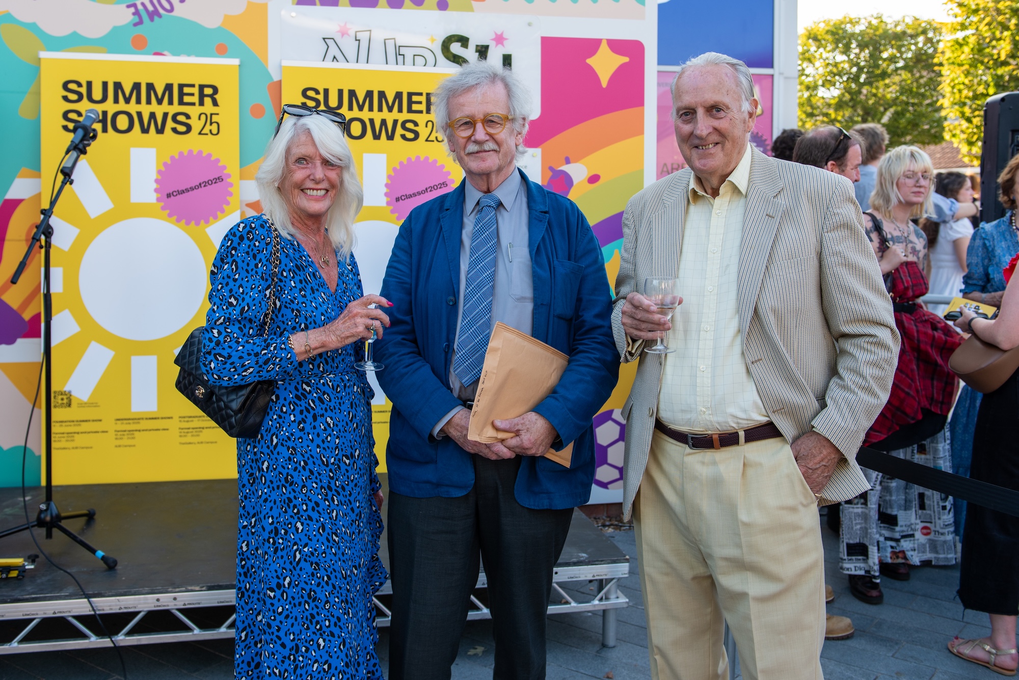 Professor Sir C. Frayling with two AUB visitors standing in front of the main stage at the 2025 Summer Shows event, with yellow Summer Shows 2025 banners in the background.