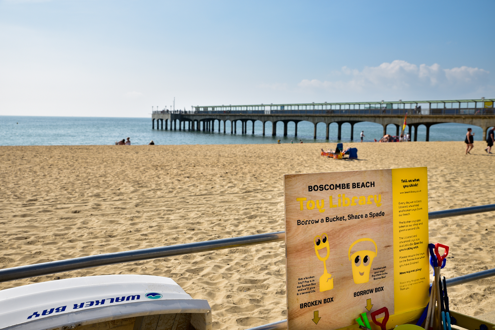 A beach with a metal railing in the foreground and a poster with text 'Boscombe Beach Toy Library' along the top. A pier is in the background.