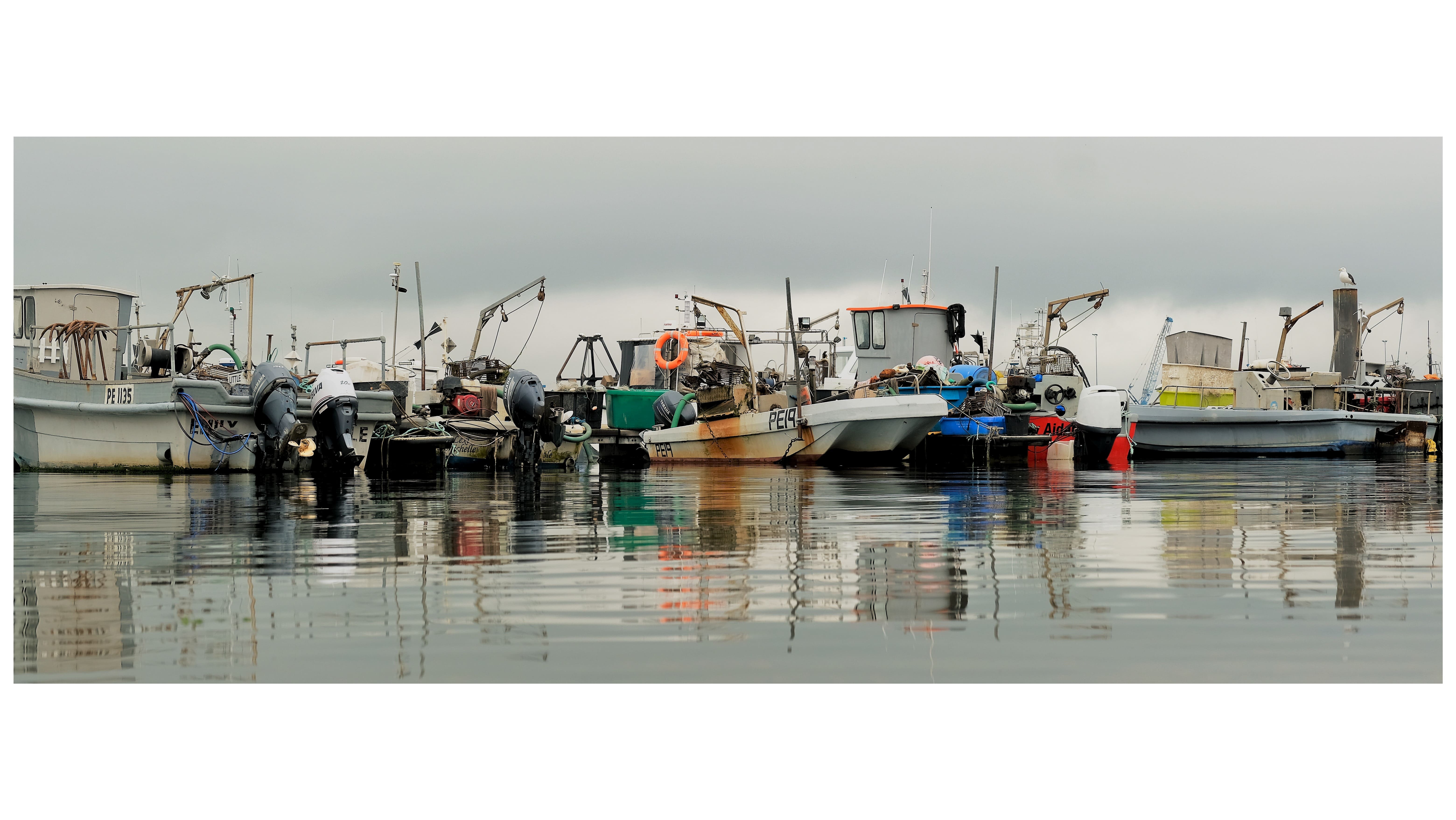 Row of colourful fishing boats moored in calm harbour water with perfect reflections beneath grey overcast sky.