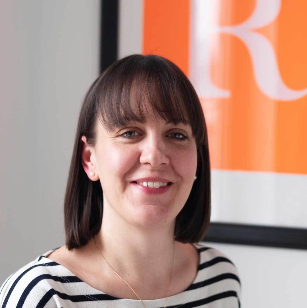 Woman with brown bob haircut and fringe wearing black and white striped top, smiling at camera with orange and white artwork behind.
