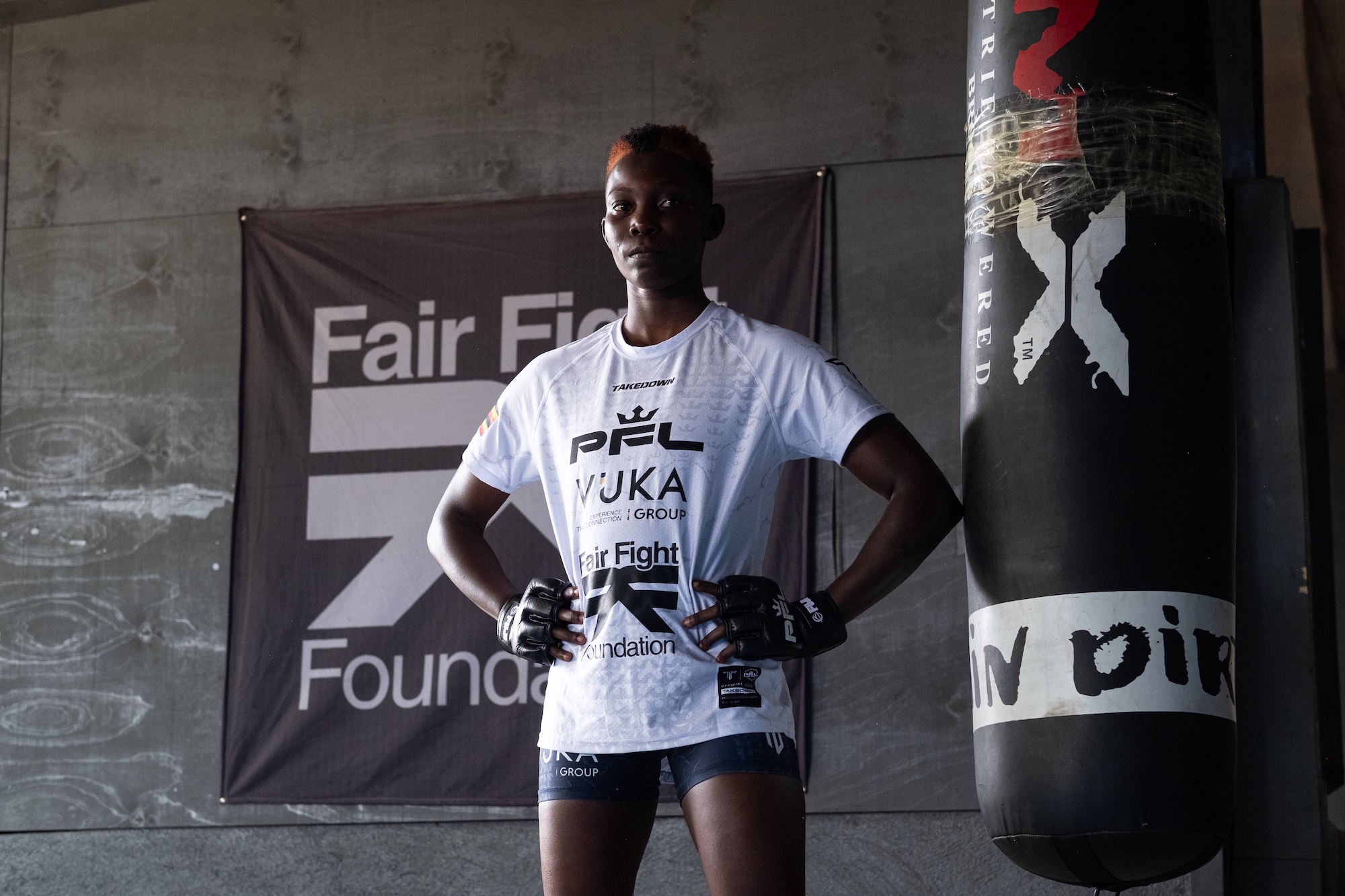 A person wearing a white T-shirt with the PFL logo stands confidently in front of a gym backdrop featuring the Fair Fight Foundation banner. A black punching bag is visible beside them.