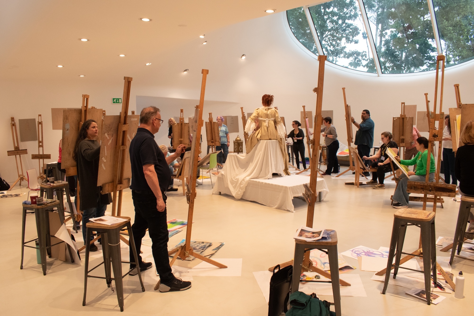 Art installation in white gallery space with vertical wooden poles on metal stands, visitors observing, circular skylight overhead.