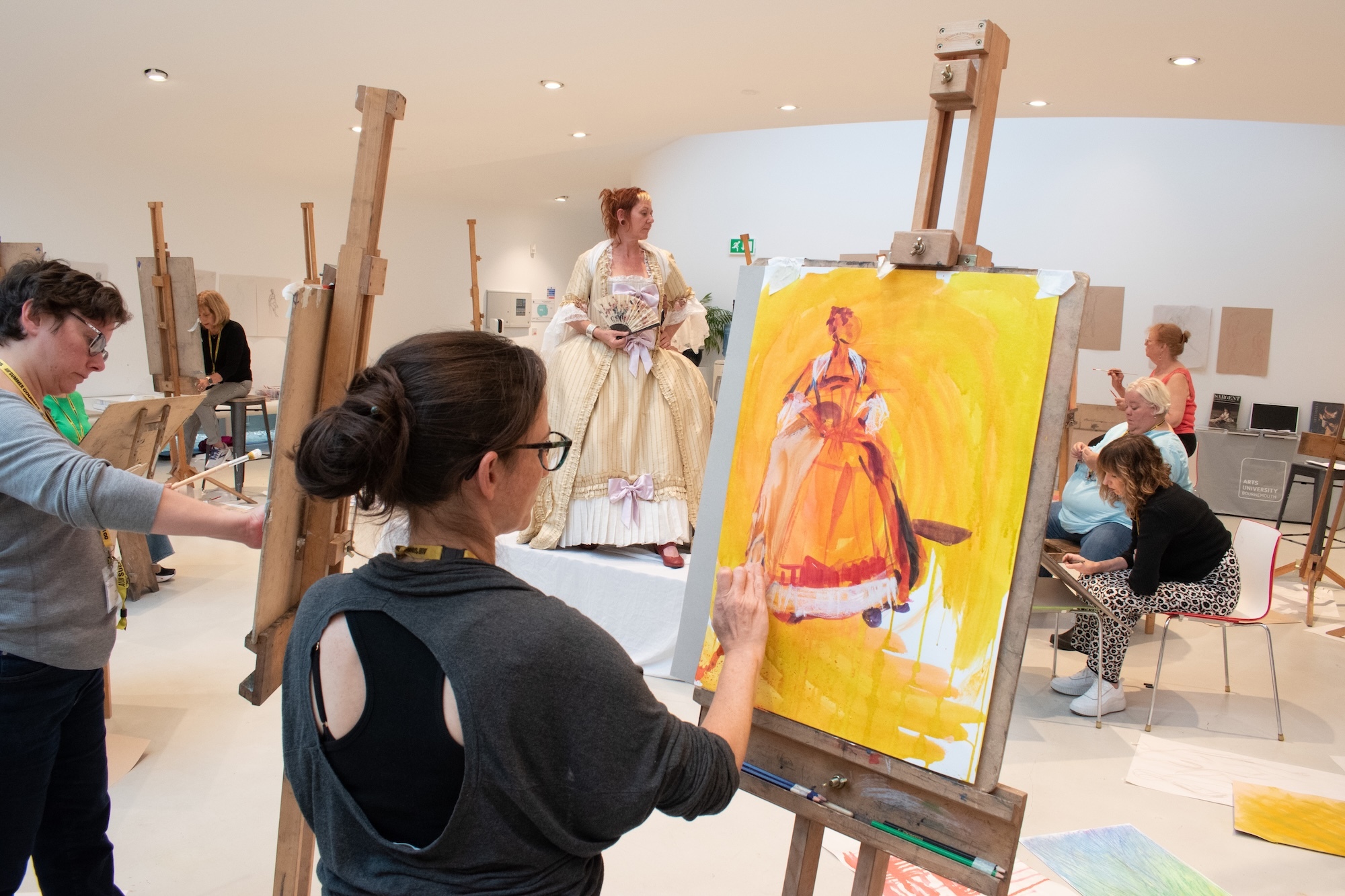 Art class with students painting at easels, woman in period dress posing as model in centre, bright studio with wooden easels and canvases.