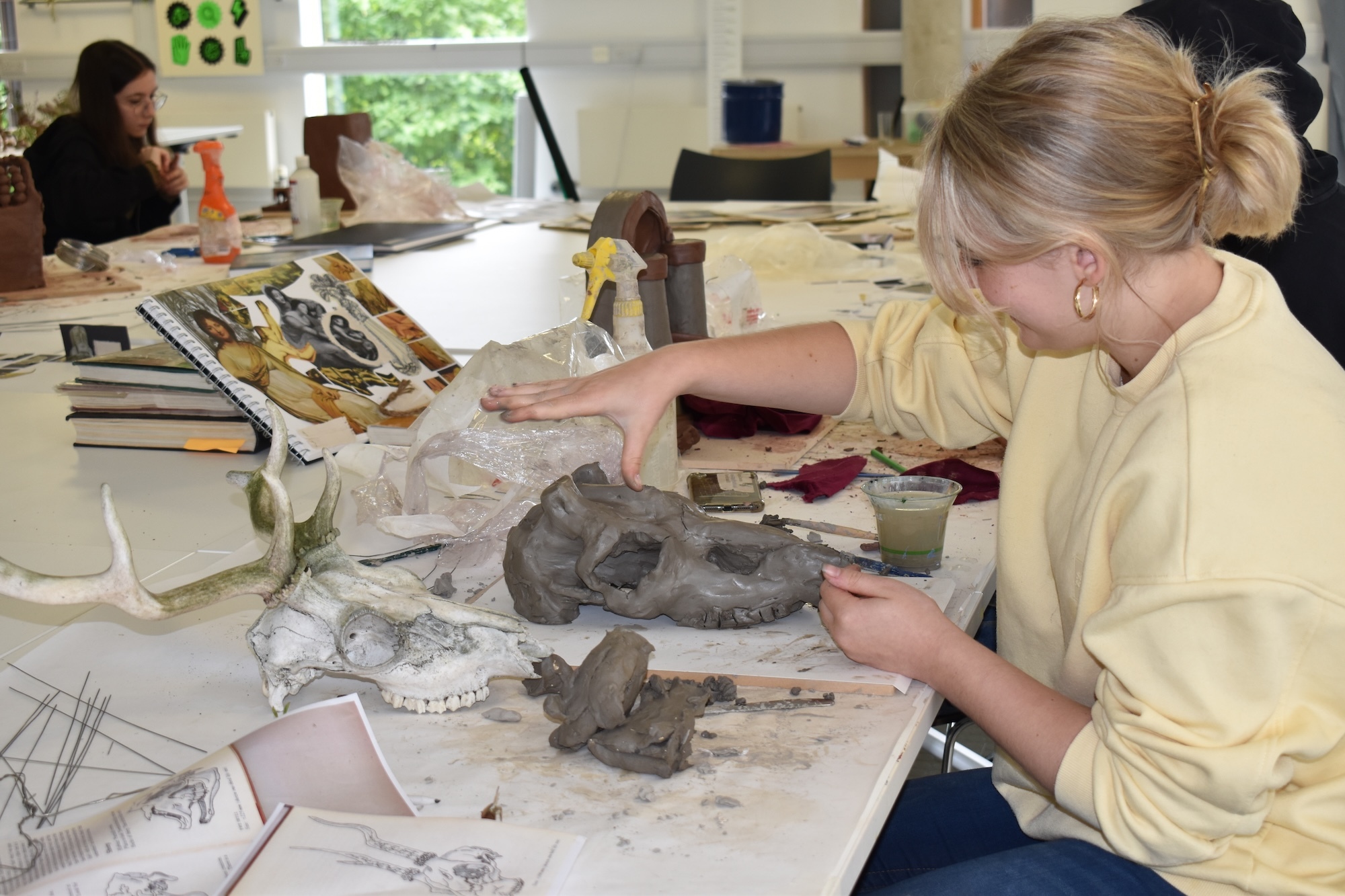 Woman in yellow top working with grey clay sculpture at table with art supplies and reference books in bright classroom setting.