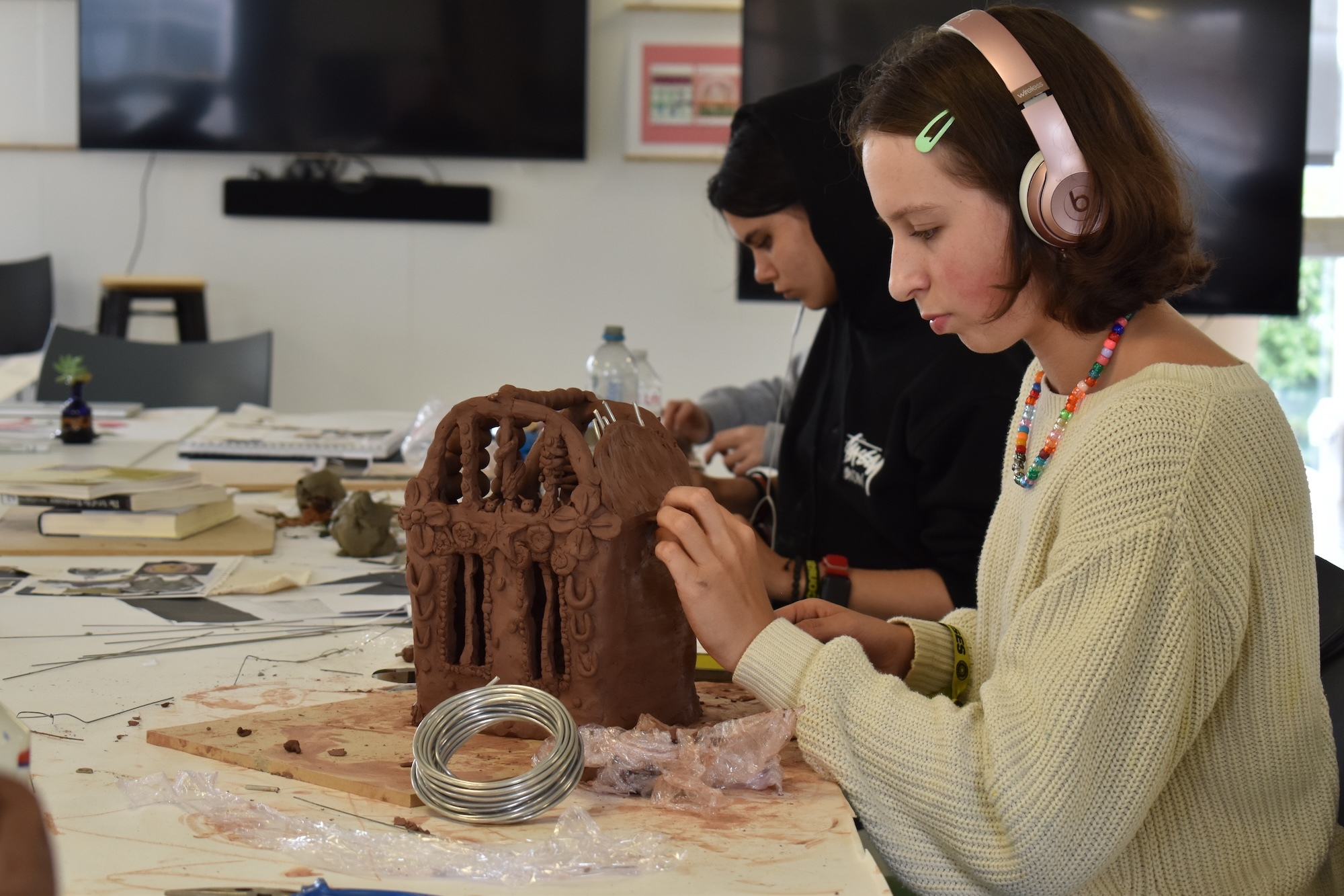 Young person in cream jumper and pink headphones working on brown clay sculpture at table with art supplies in workshop setting