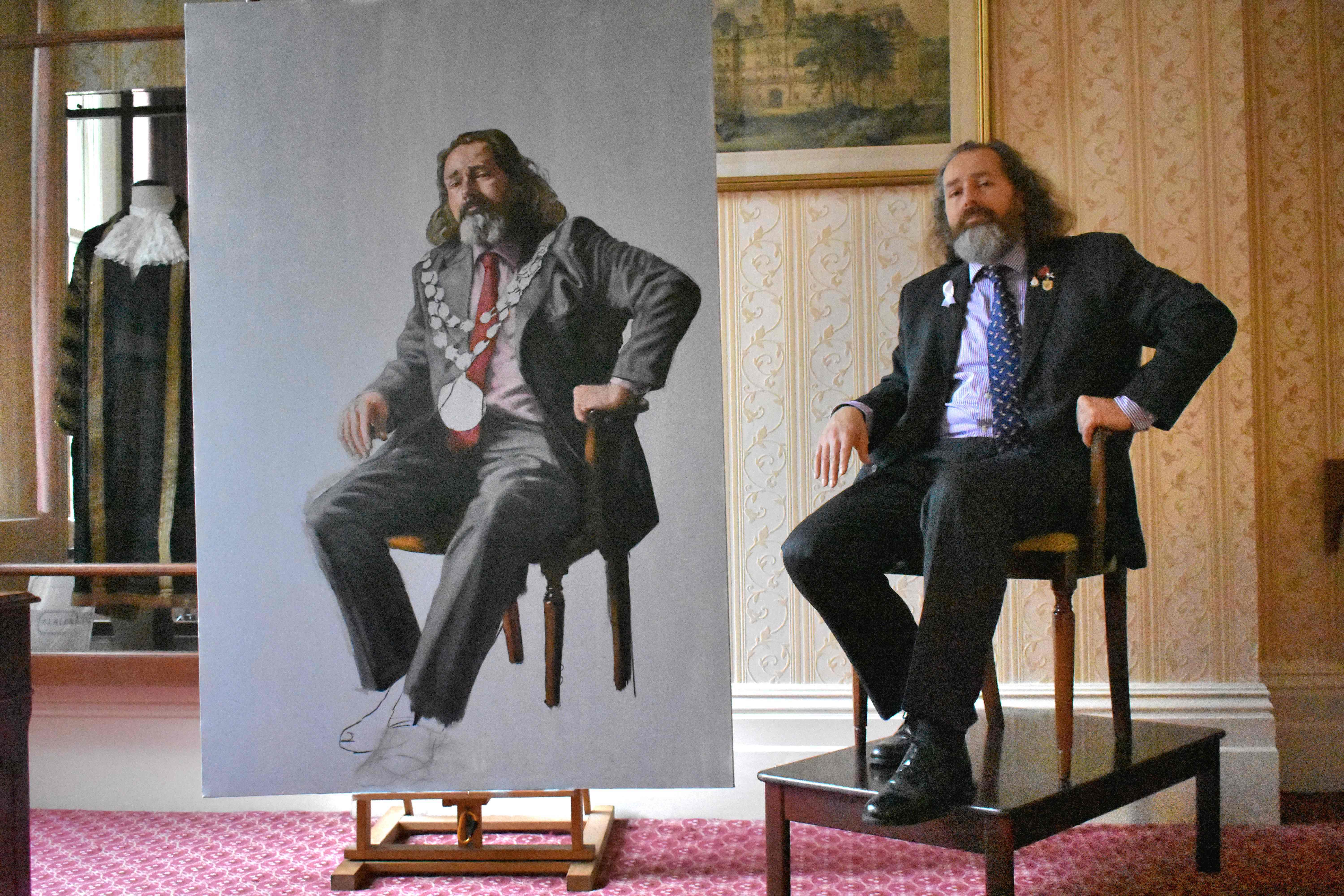 Bearded man in dark suit sits on chair beside large portrait painting of similarly posed bearded figure, ornate room with patterned wallpaper.