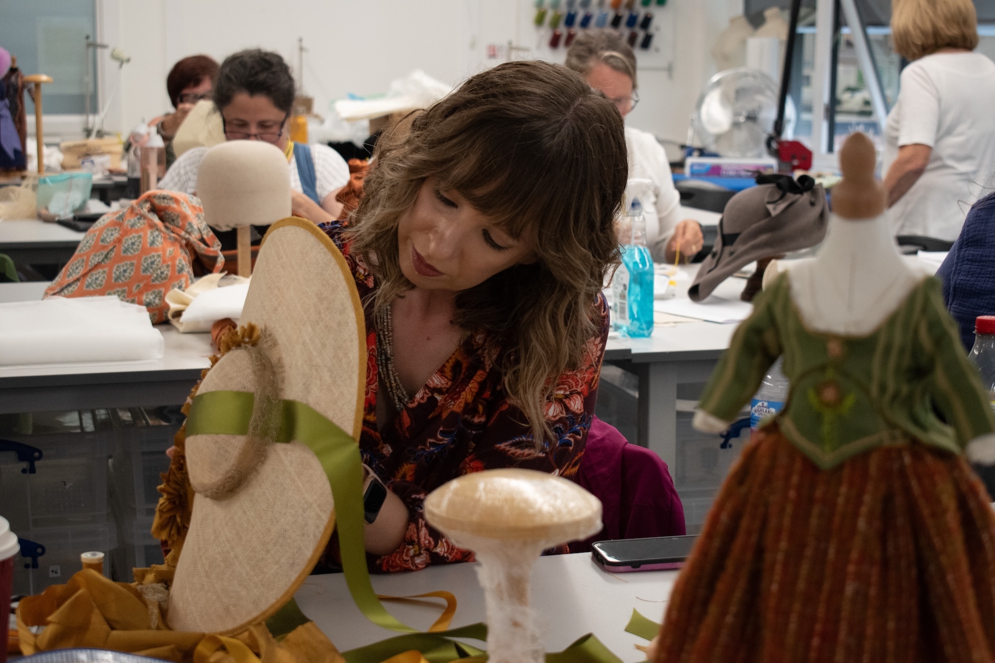 Woman with brown hair working on white dress form in busy sewing workshop with multiple people at tables in background.